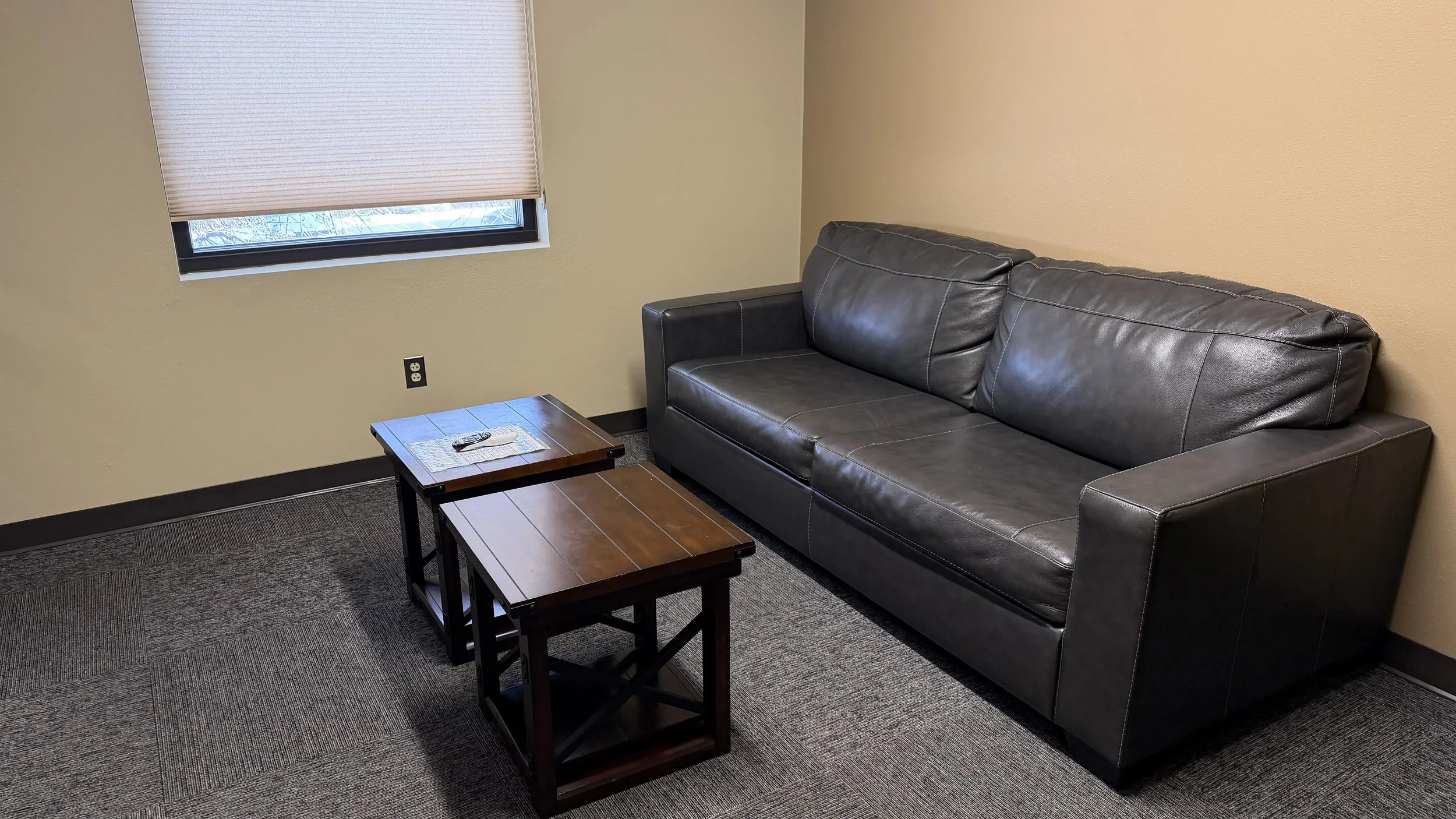 A small office or waiting room with a leather couch, two wooden tables, a window with closed blinds, a beige wall, and an electrical outlet.