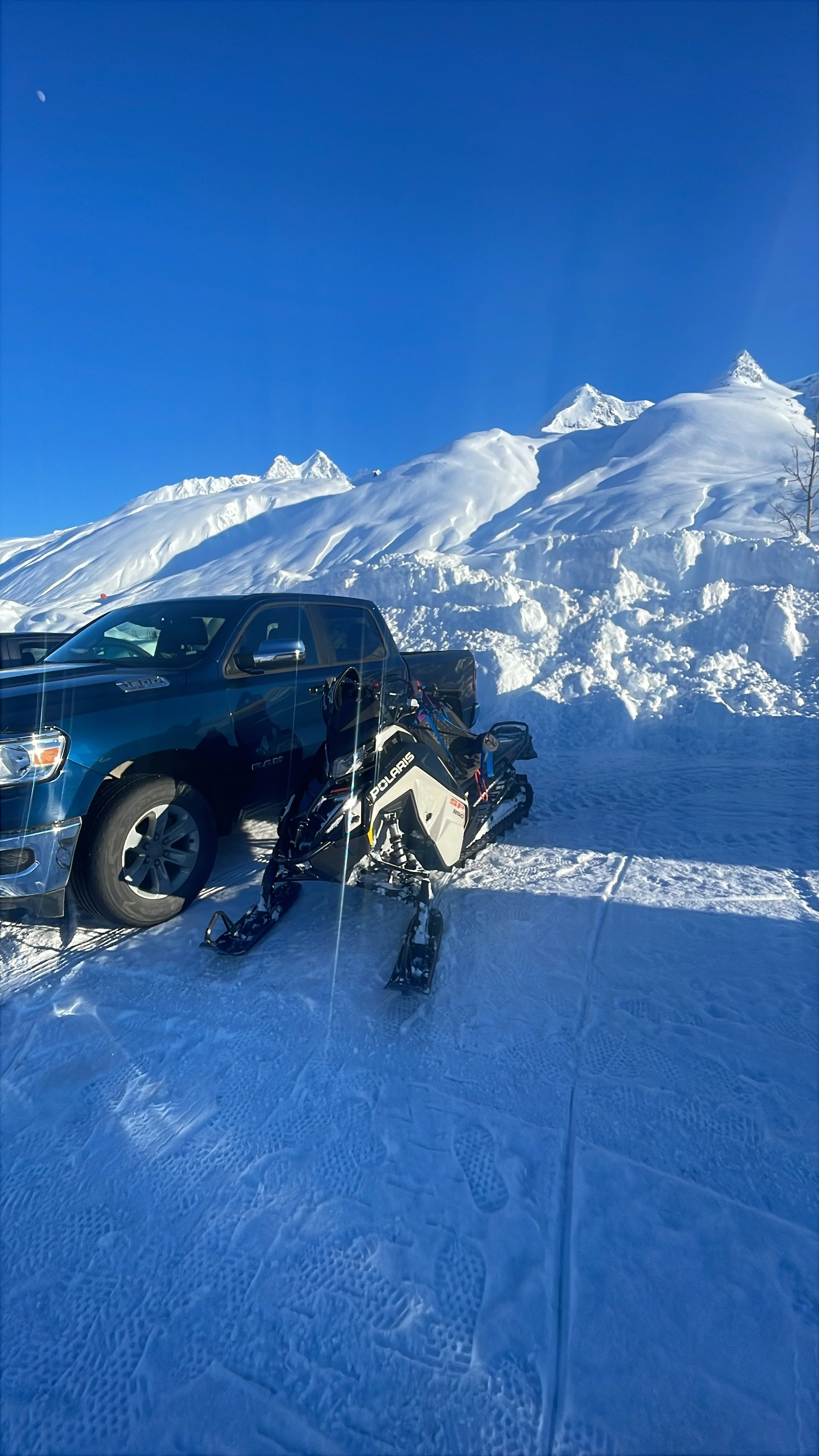 A snow-covered landscape with a blue truck and a new Polaris snowmobile in front of snowy mountains under a clear blue sky.