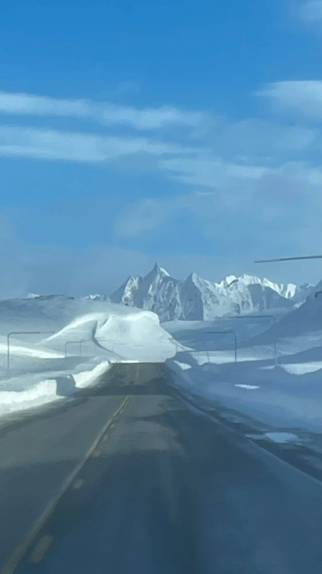 Snow-covered mountain range seen through a car windshield during daylight with the road blocked by snow drift.