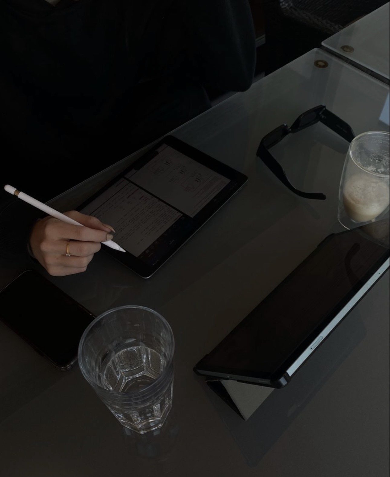 Person using a stylus on a tablet, with glasses, a phone, and drinks on the table.