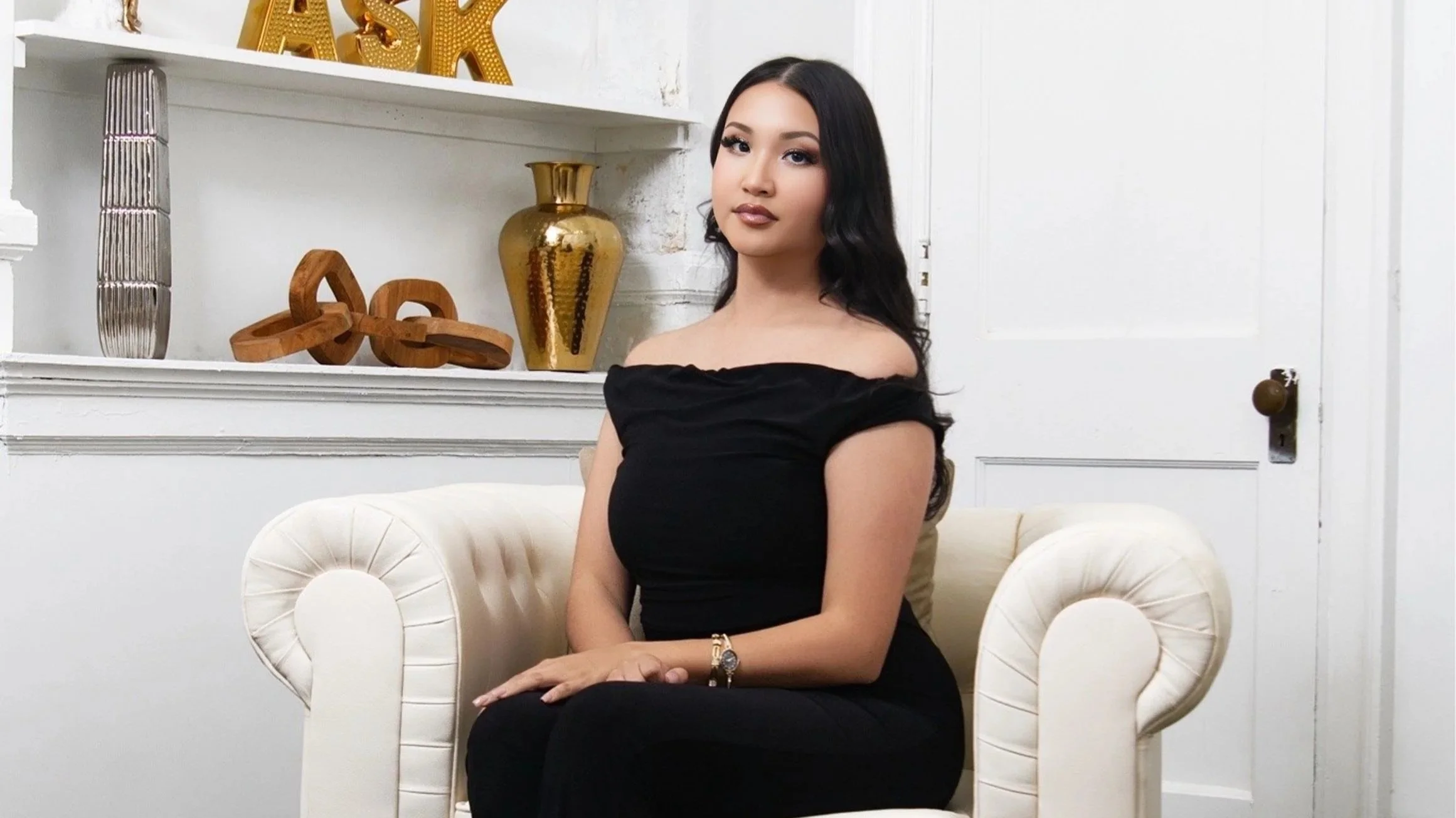 A woman with long black hair, wearing a black off-shoulder dress, is sitting on a cream-colored tufted armchair in a decorated room with white walls. Decorative items, including a gold vase, wooden sculptures, and large gold letters, are on a shelf behind her.