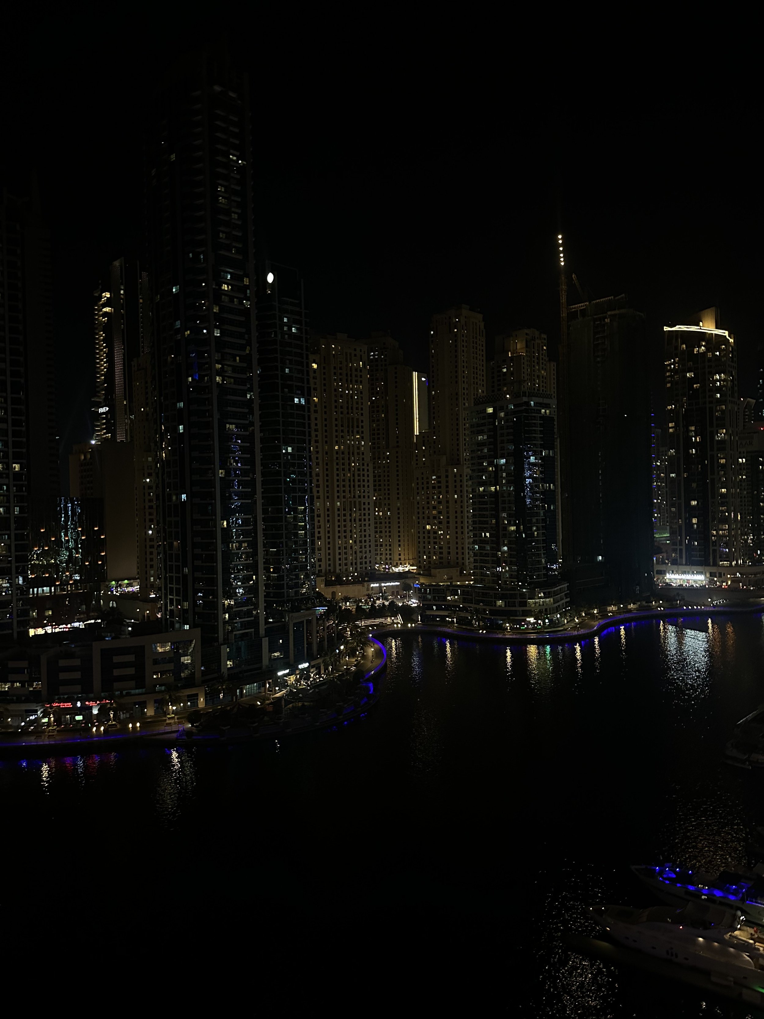 Nighttime view of a city skyline with tall illuminated buildings reflected in a body of water, featuring some boats and a dark sky.