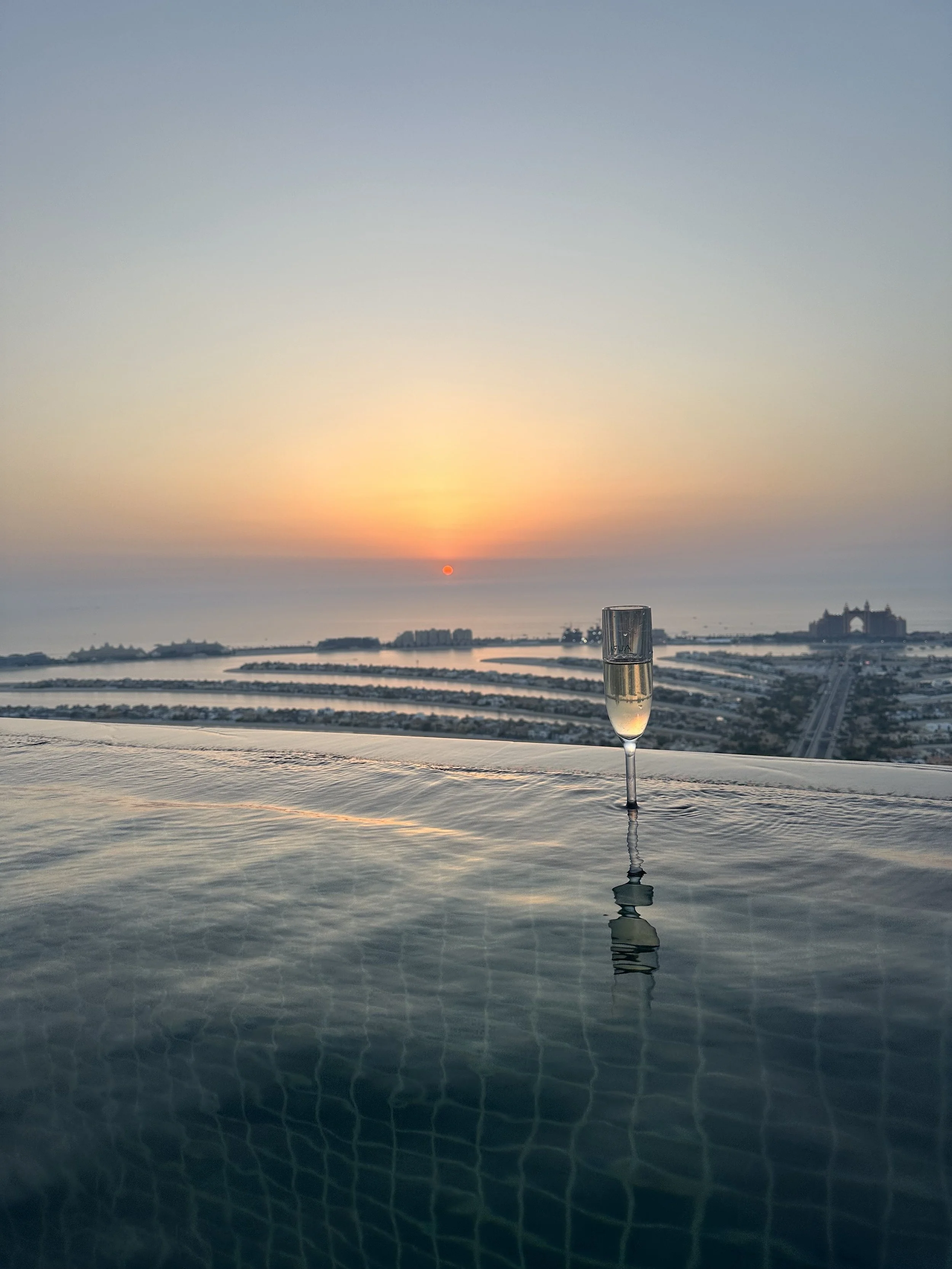 A glass of champagne on an infinity pool with a sunset view over a city and water in the background.
