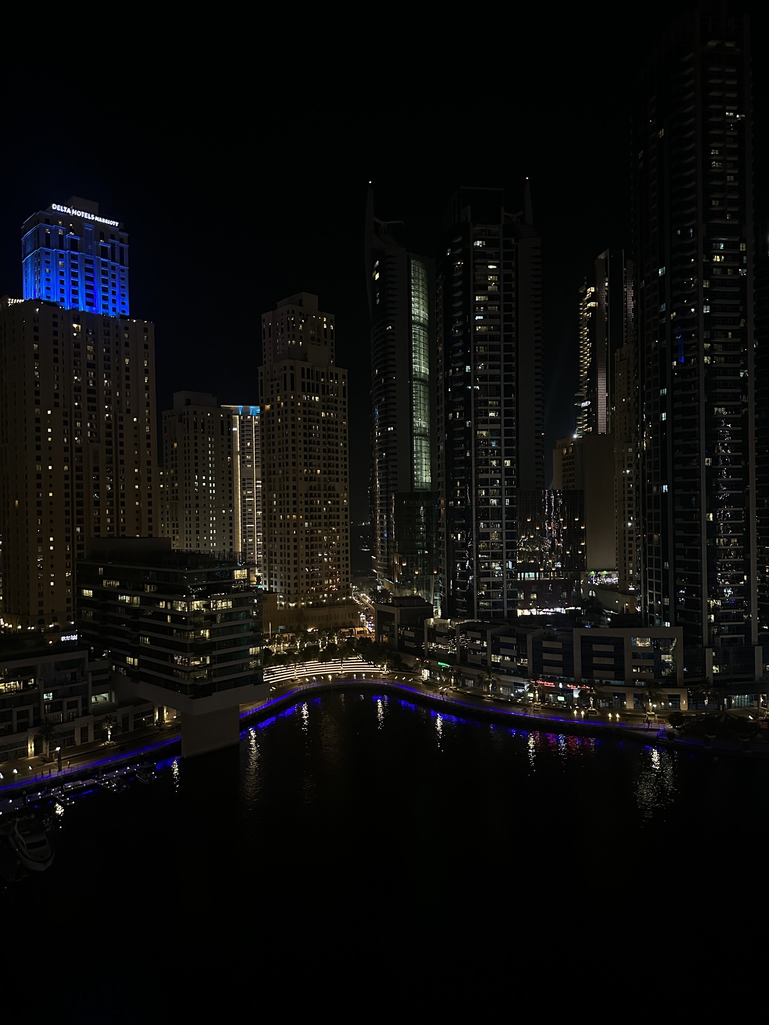 Nighttime view of a city skyline with illuminated tall skyscrapers and a waterway reflecting blue and white lights.