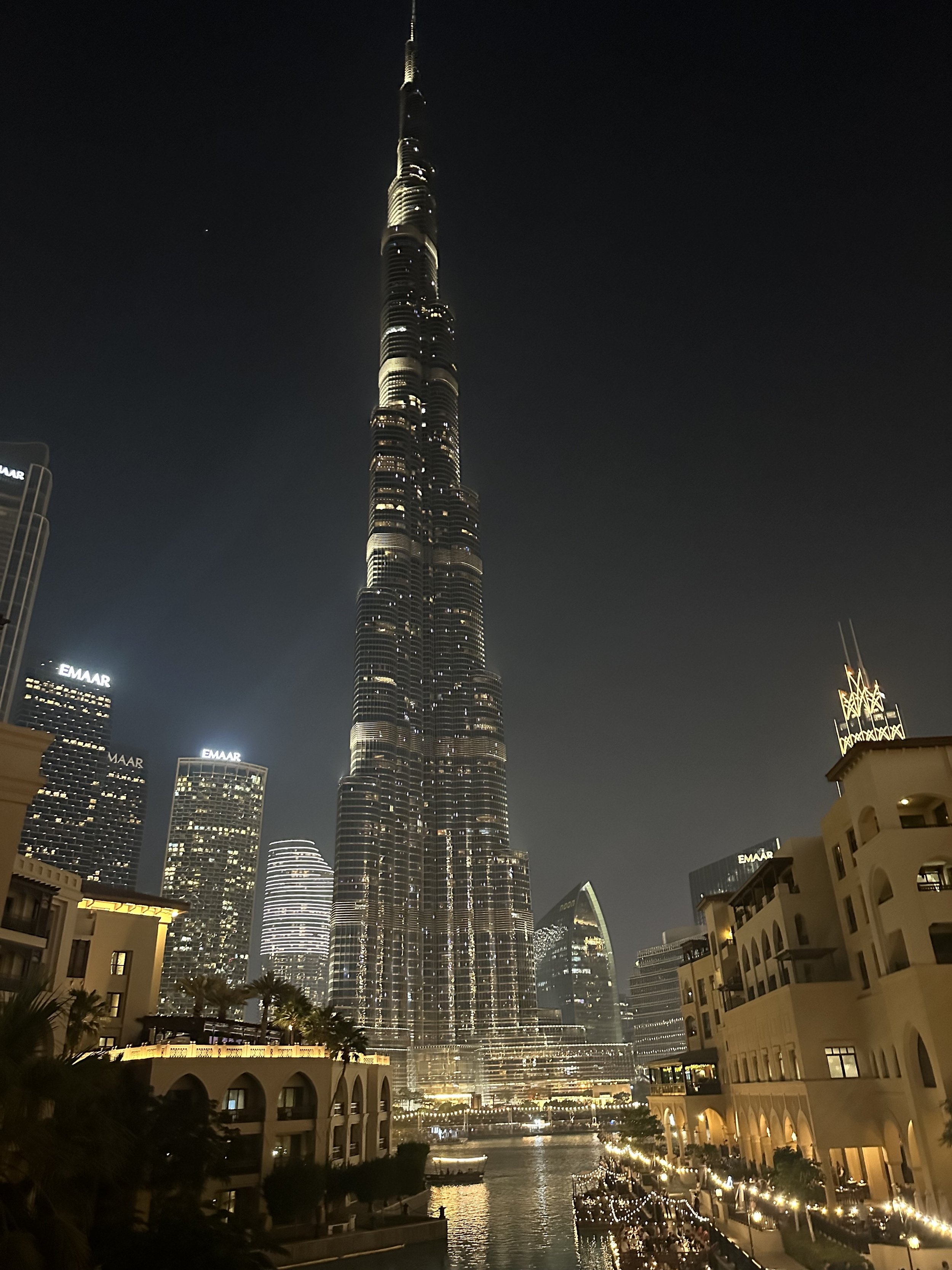 Night view of the Burj Khalifa skyscraper in Dubai, illuminated with lights, with surrounding buildings and a canal in the foreground.