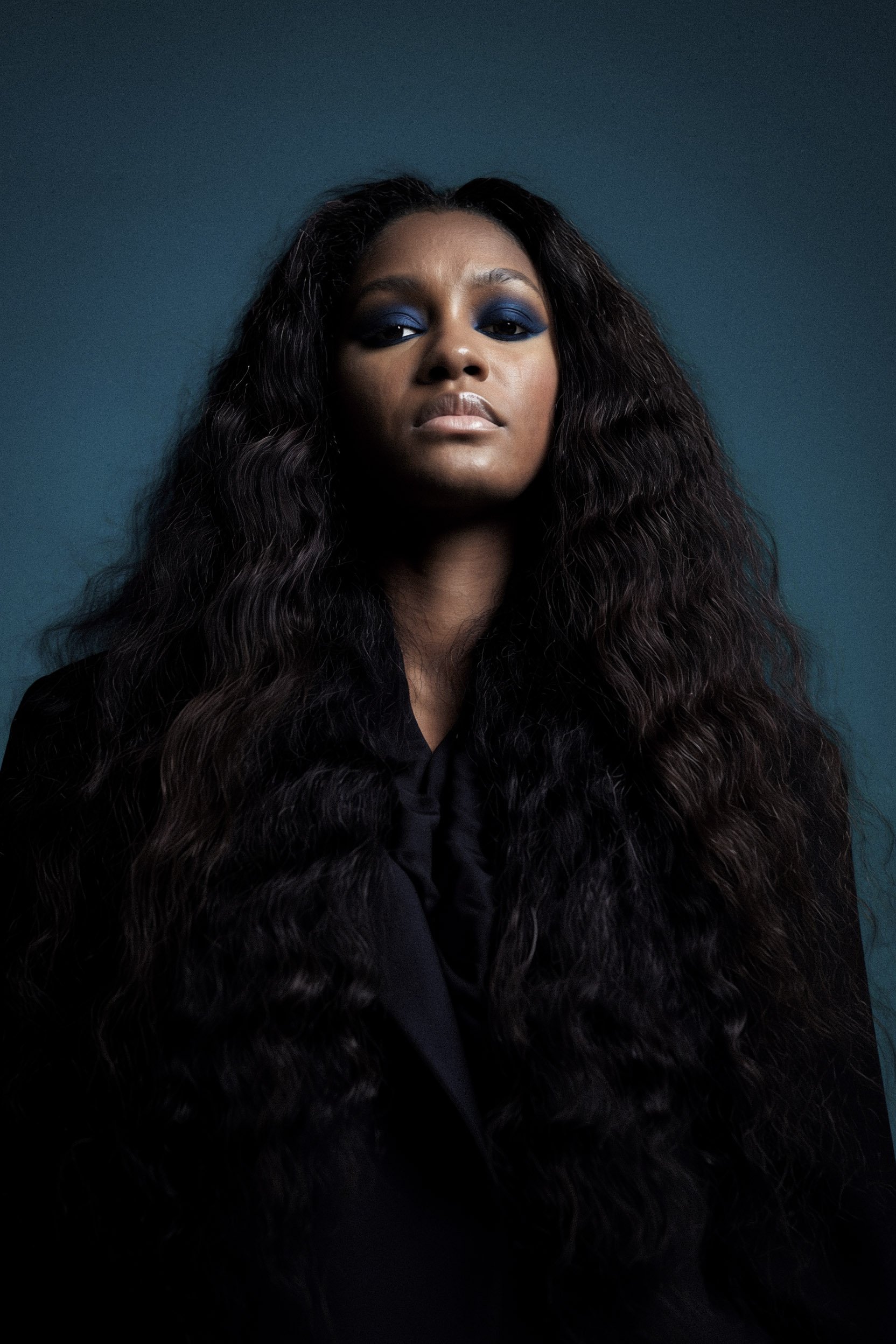 Portrait of a woman with long, curly dark hair, wearing a black top and blue eyeshadow, against a dark teal background.