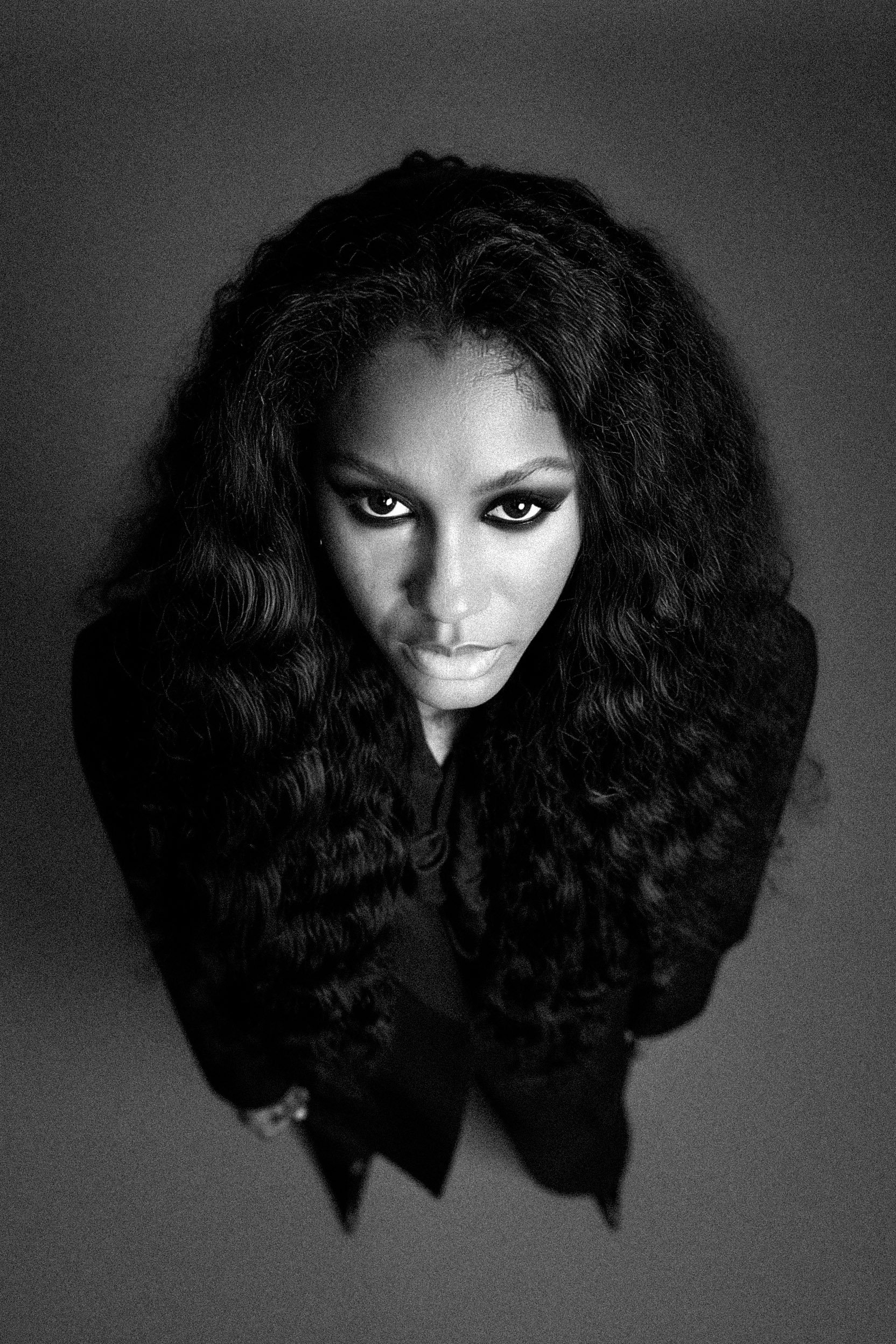 Black and white portrait of a woman with long, curly hair, looking up at the camera with intense eyes.