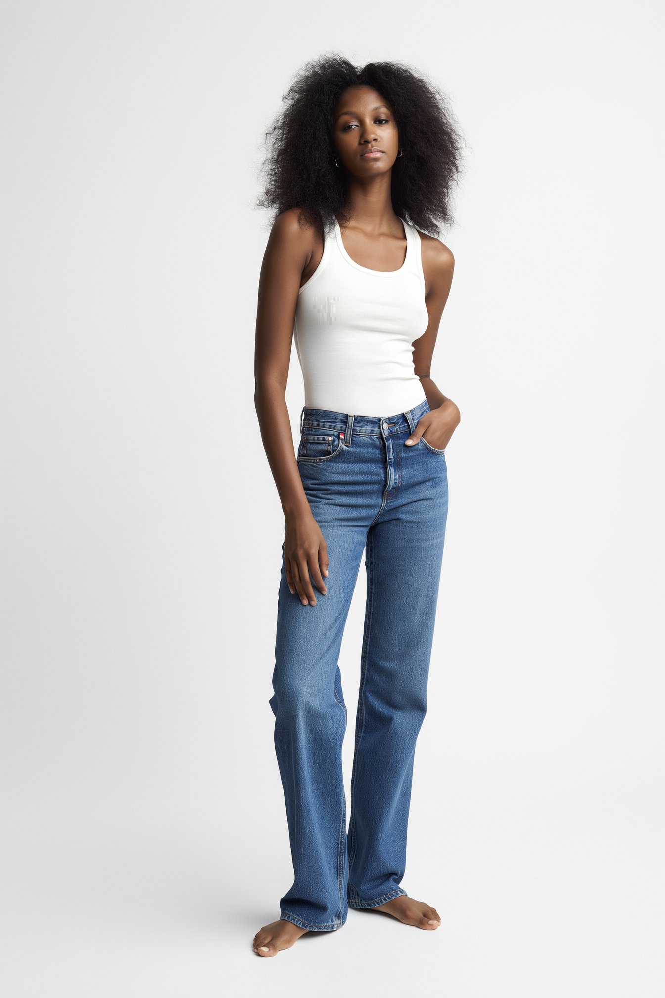 A young woman with curly hair wearing a white tank top and blue jeans standing barefoot against a white background.