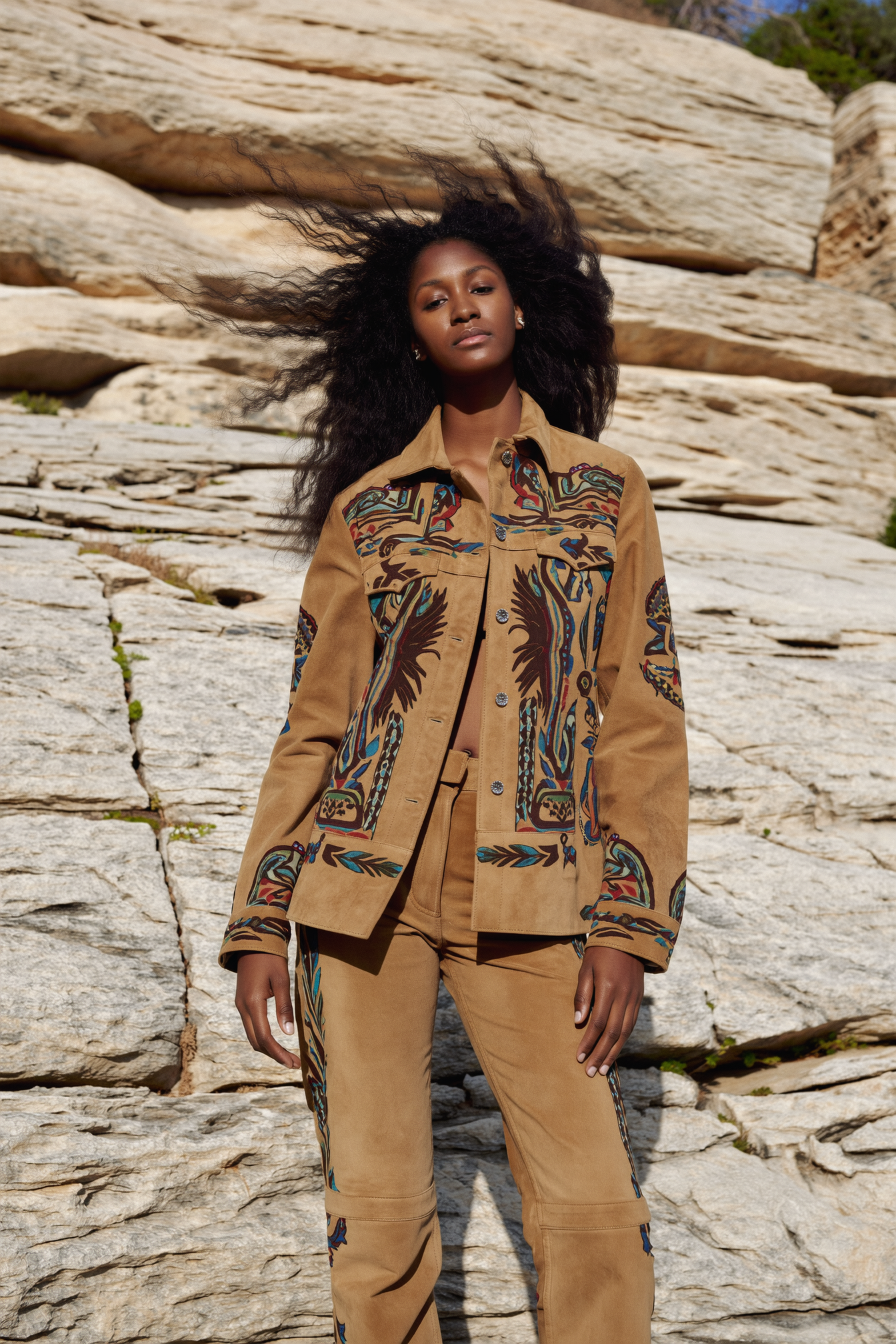 A woman with long, curly hair stands outdoors against a rocky background, wearing a tan vintage-style embroidered jacket and matching pants.