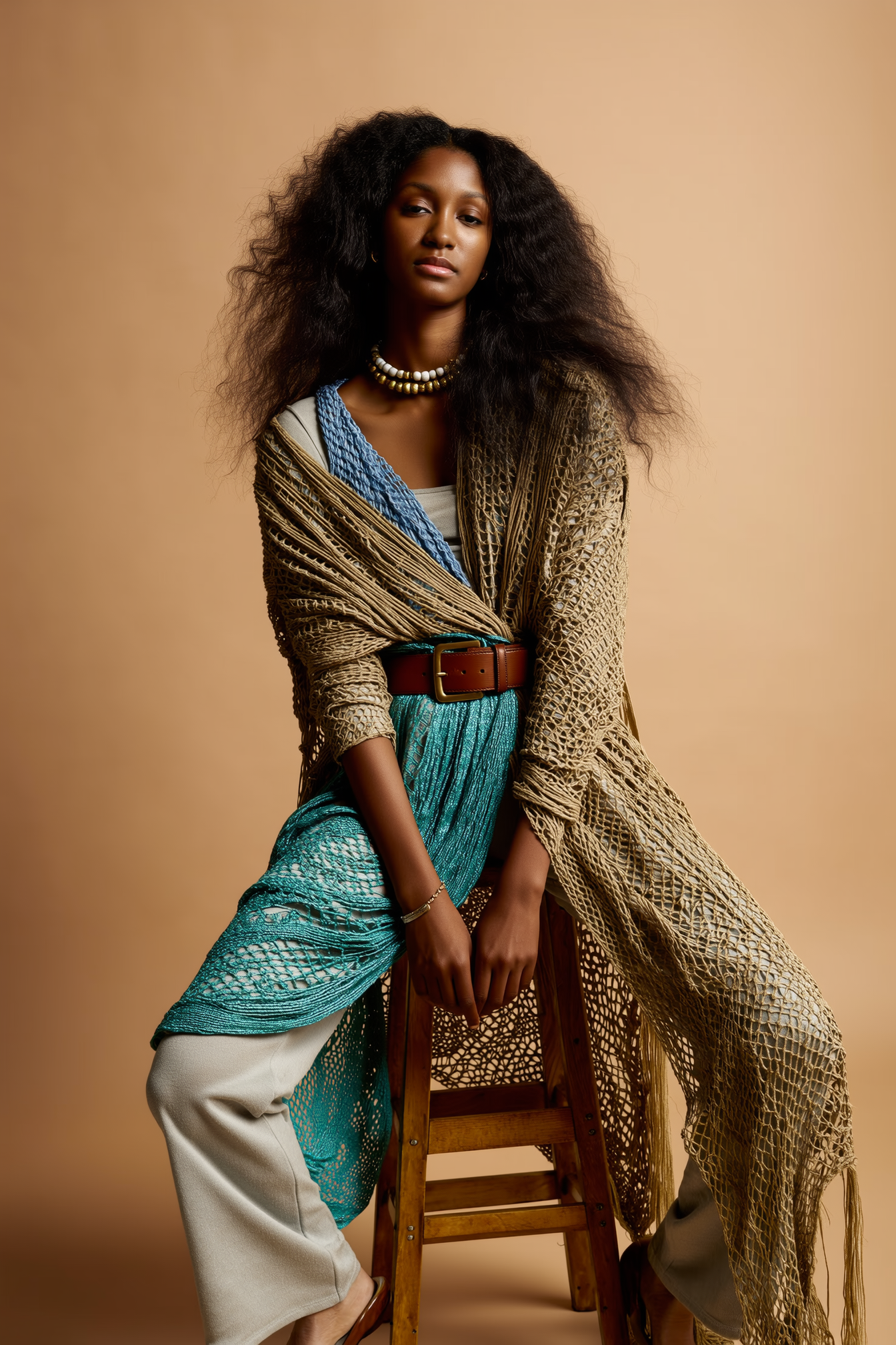 A woman with voluminous curly hair wearing a layered outfit with a crochet cardigan, a blue top, cream-colored pants, and accessorized with necklaces and a brown belt, sitting on a wooden stool against a beige background.
