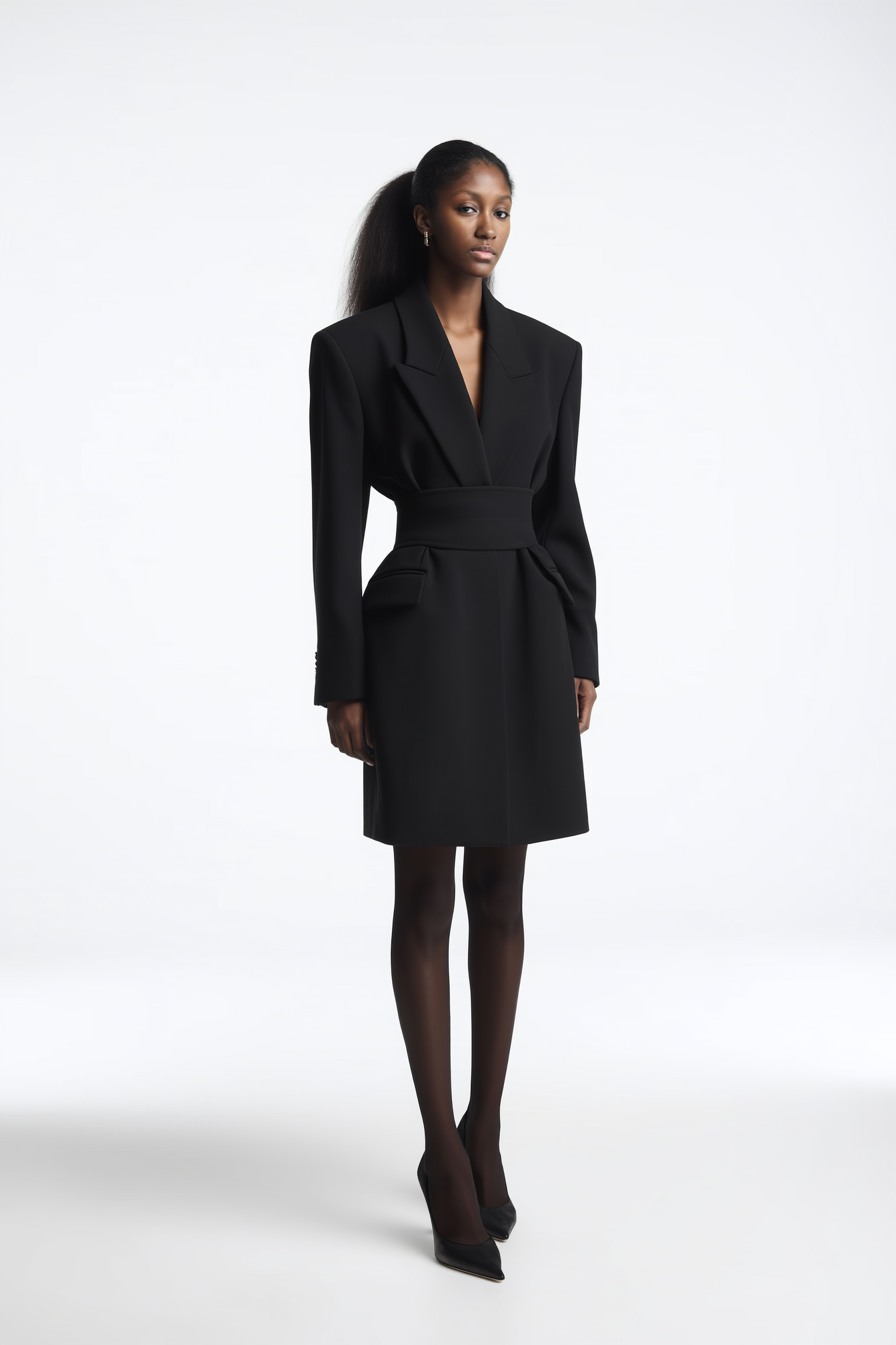 A woman in a black blazer dress, black stockings, and black heels standing against a white background.