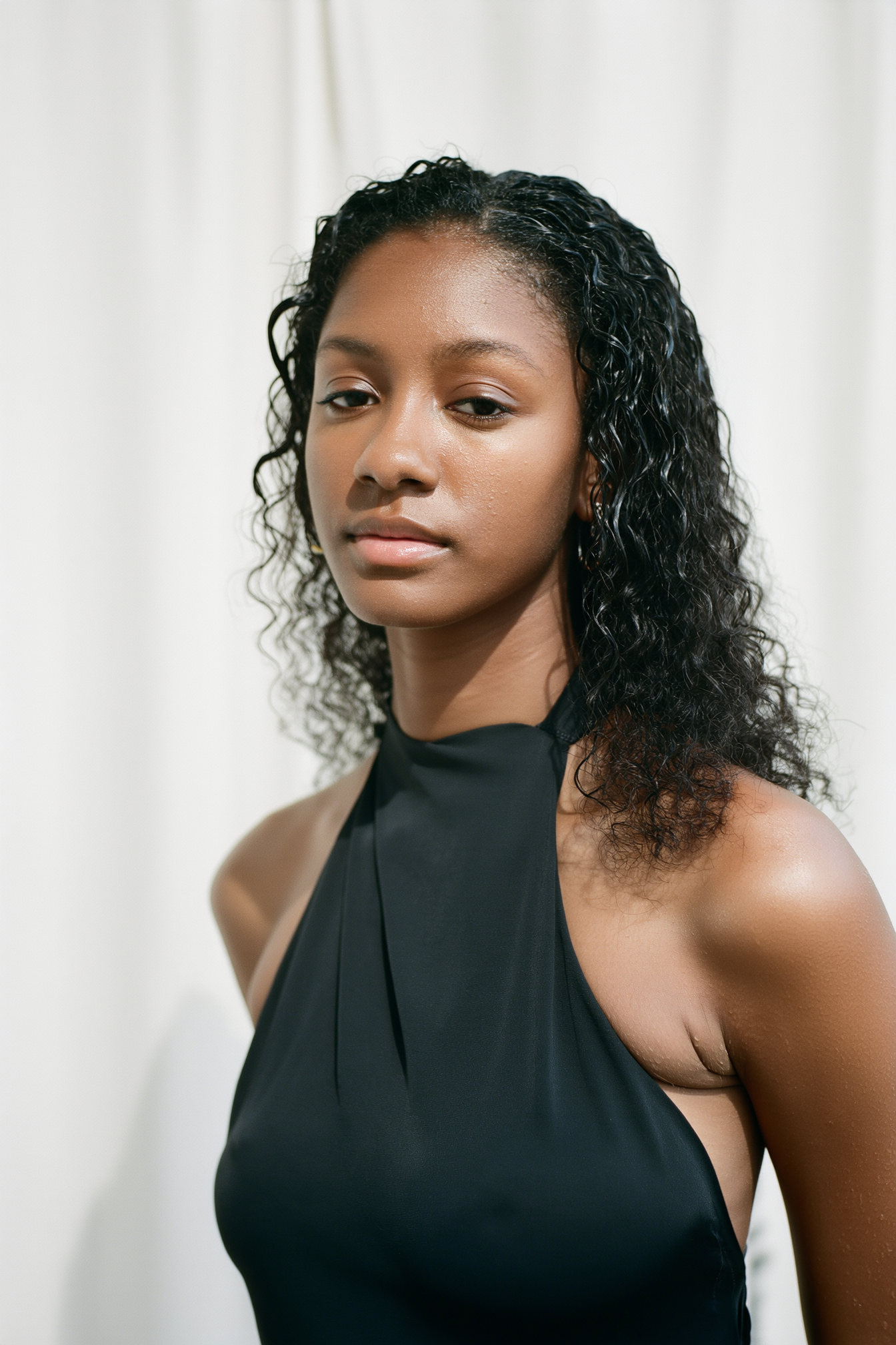 A portrait of a young woman with curly black hair wearing a sleeveless black high-neck top, standing against a plain white background.