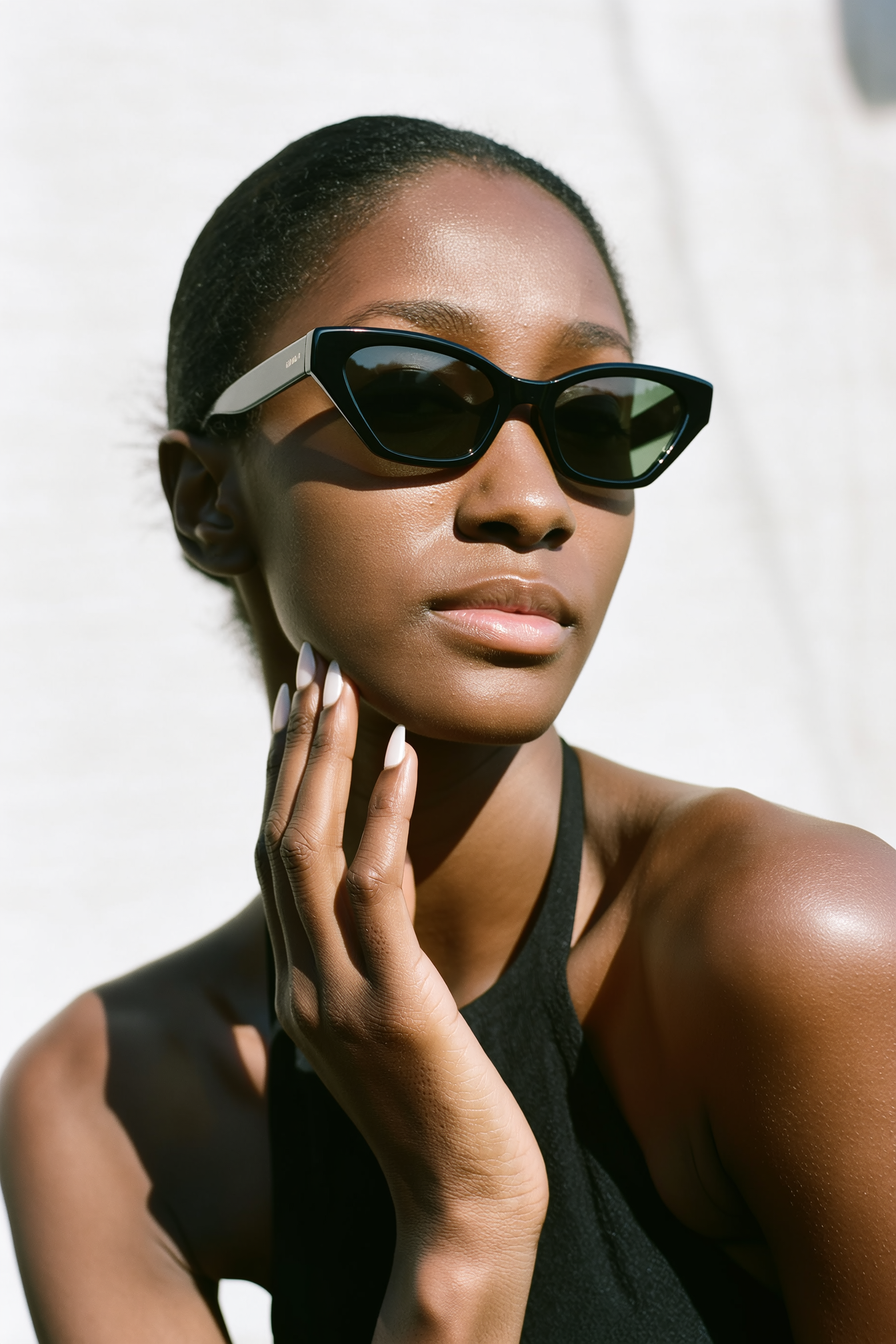 A woman with dark skin wearing black sunglasses, a black halter top, and white nail polish, touching her face with her right hand.