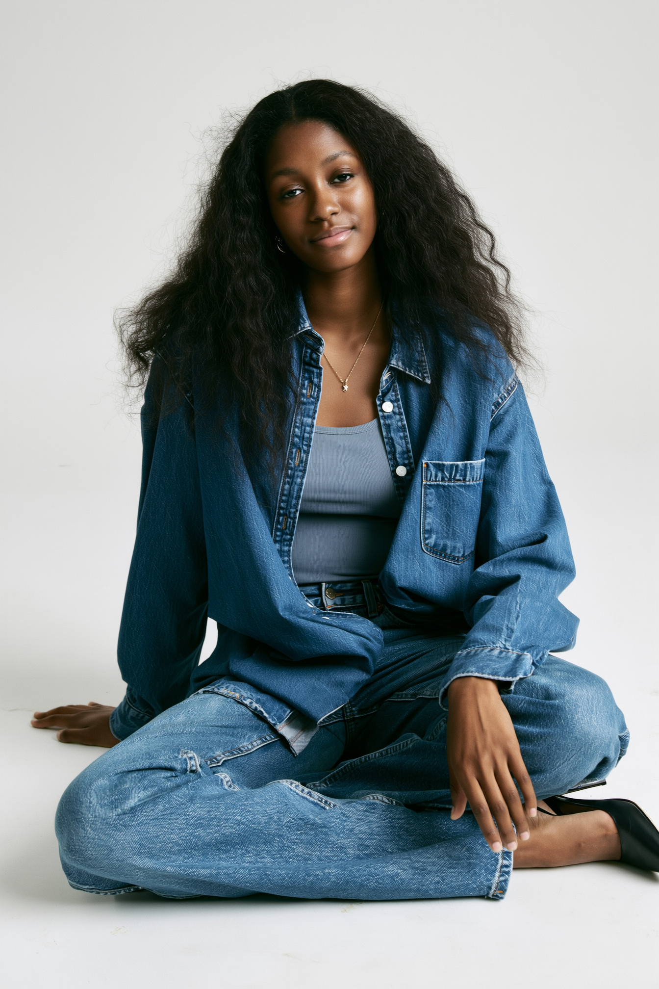 A young woman with long, curly black hair sitting cross-legged on the floor, wearing a denim jacket and jeans, with a light blue tank top underneath, and black shoes. She has a calm expression and is in front of a plain white background.