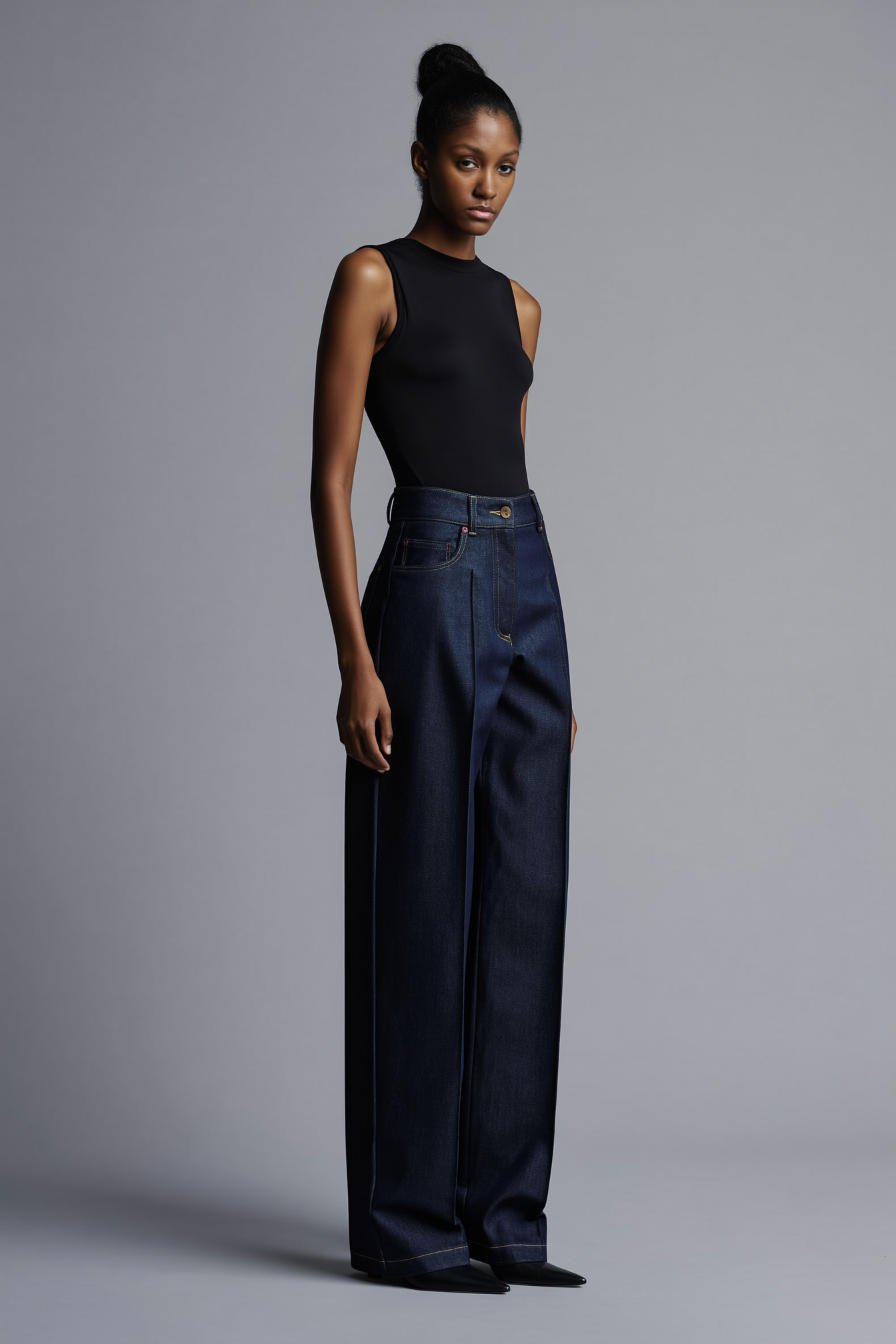 A young woman with a high bun hairstyle wearing a sleeveless black top, wide-legged dark denim jeans, and black shoes, standing against a plain gray background.