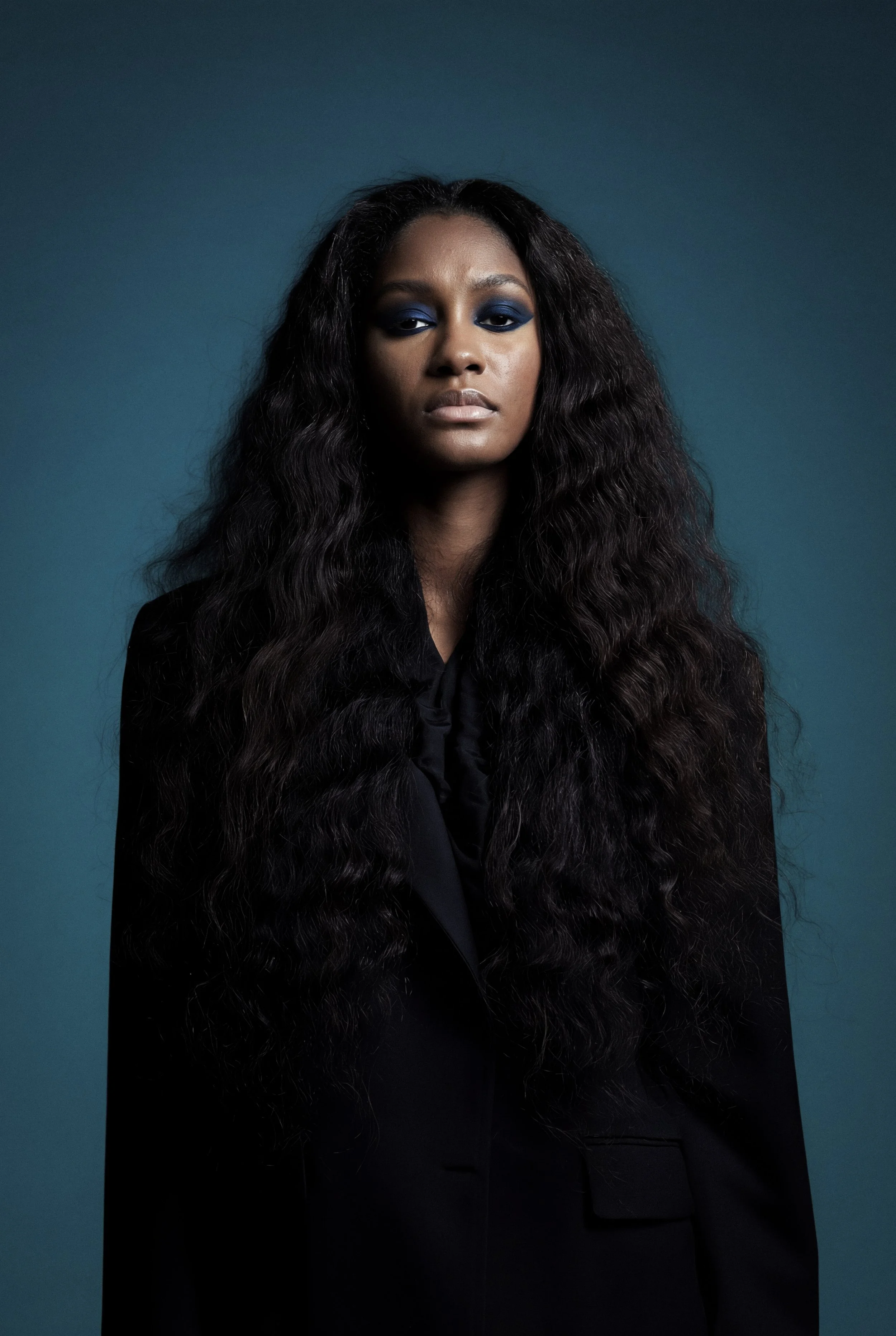 A woman with long, wavy black hair wearing a black blazer against a dark teal background.
