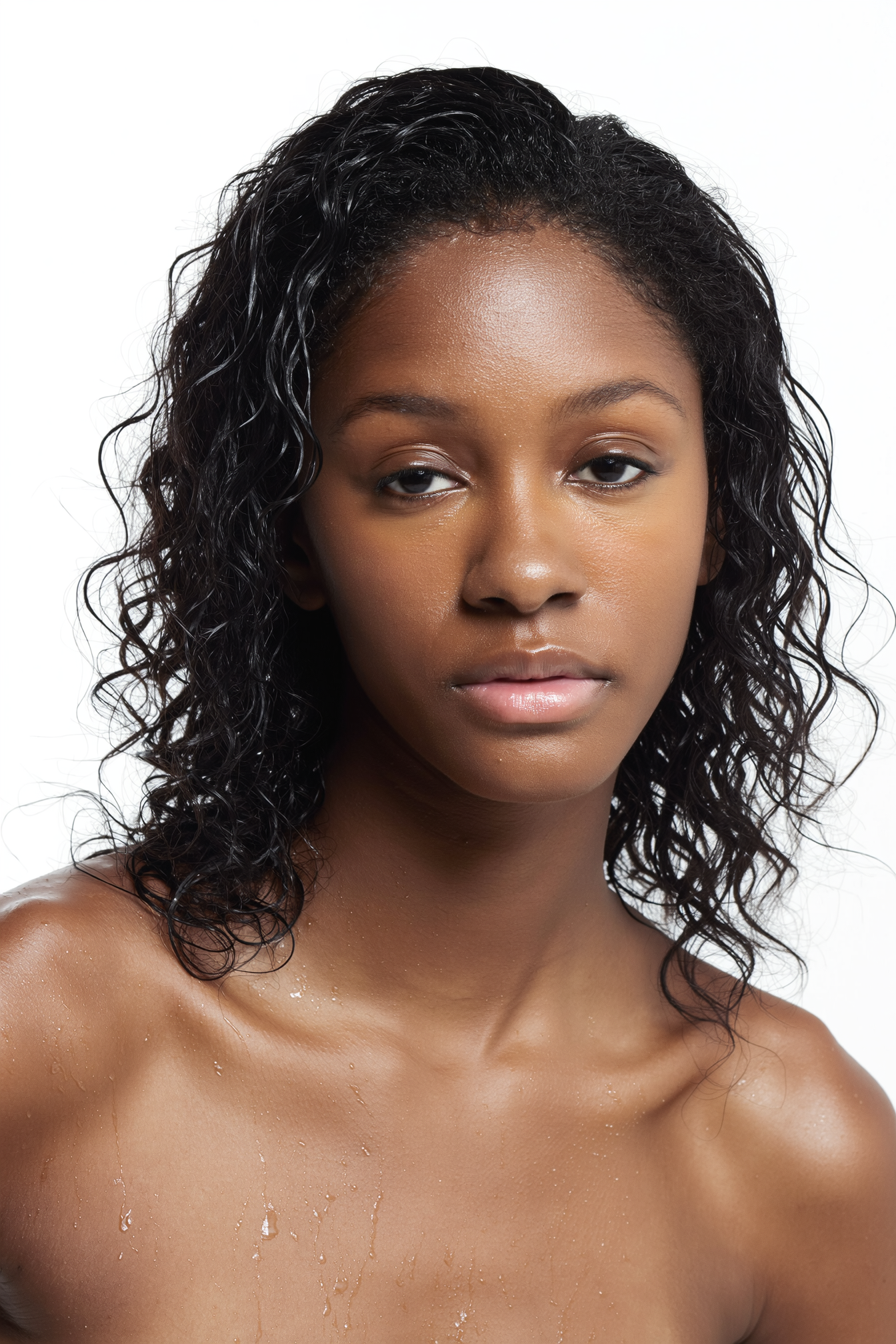 Portrait of a young woman with curly black hair and smooth brown skin, looking at the camera with a neutral expression, against a plain white background.