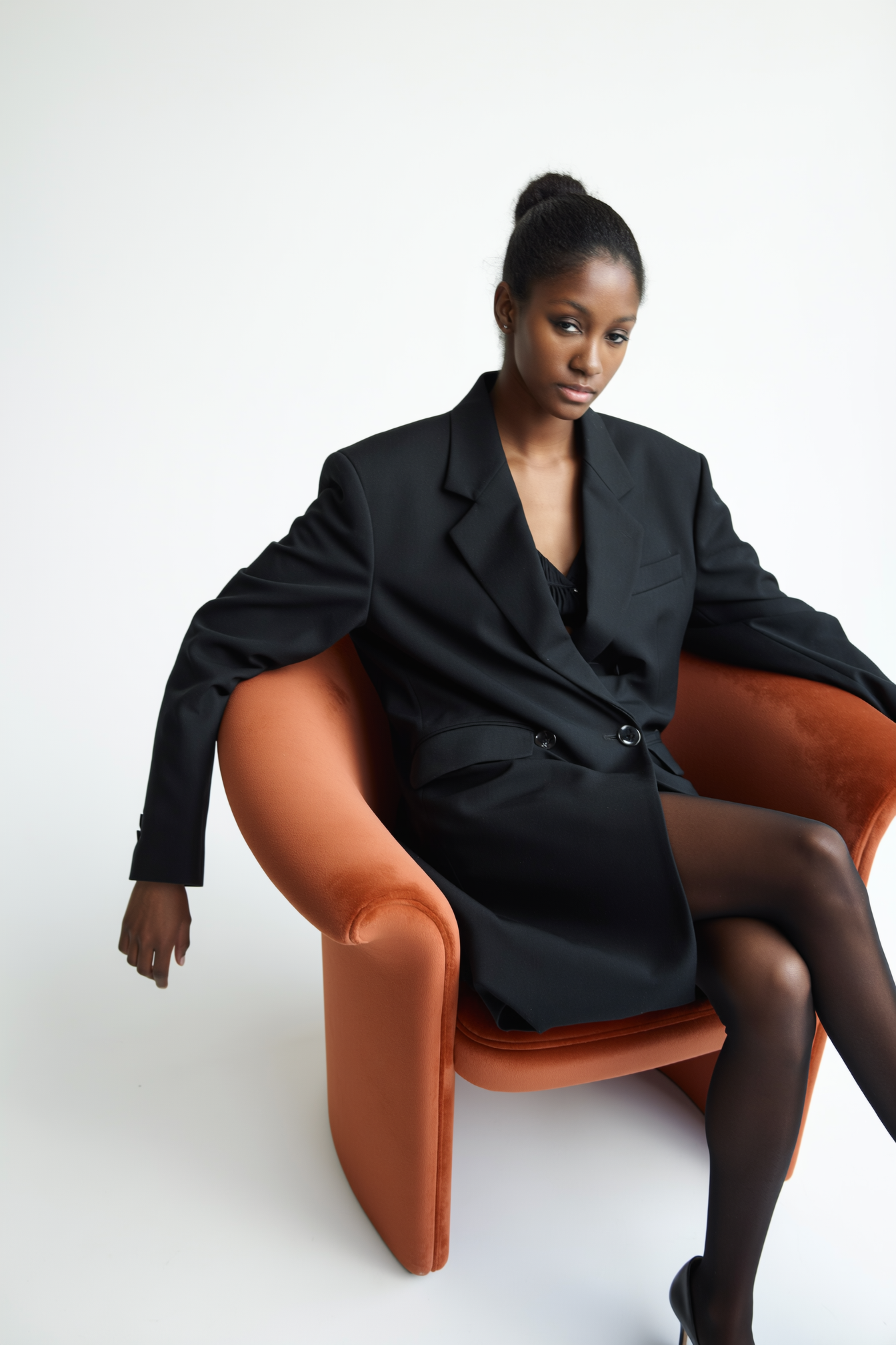 A woman wearing a black blazer and black sheer tights sitting on an orange velvet chair against a plain white background.