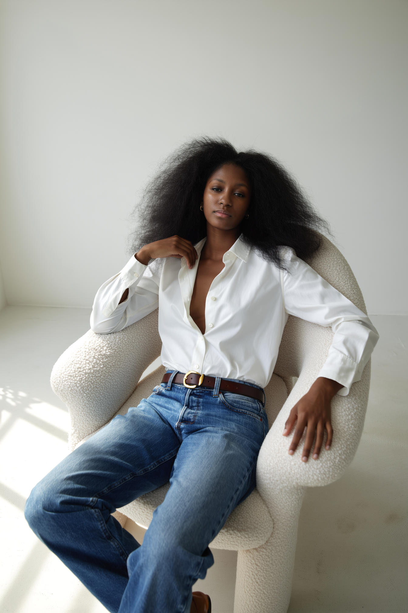 A woman with voluminous black hair sitting in a cream-colored textured armchair in a minimalist room. She is wearing a white button-up shirt with the top few buttons open, blue jeans, and a brown belt. She has a calm, confident expression and is slig