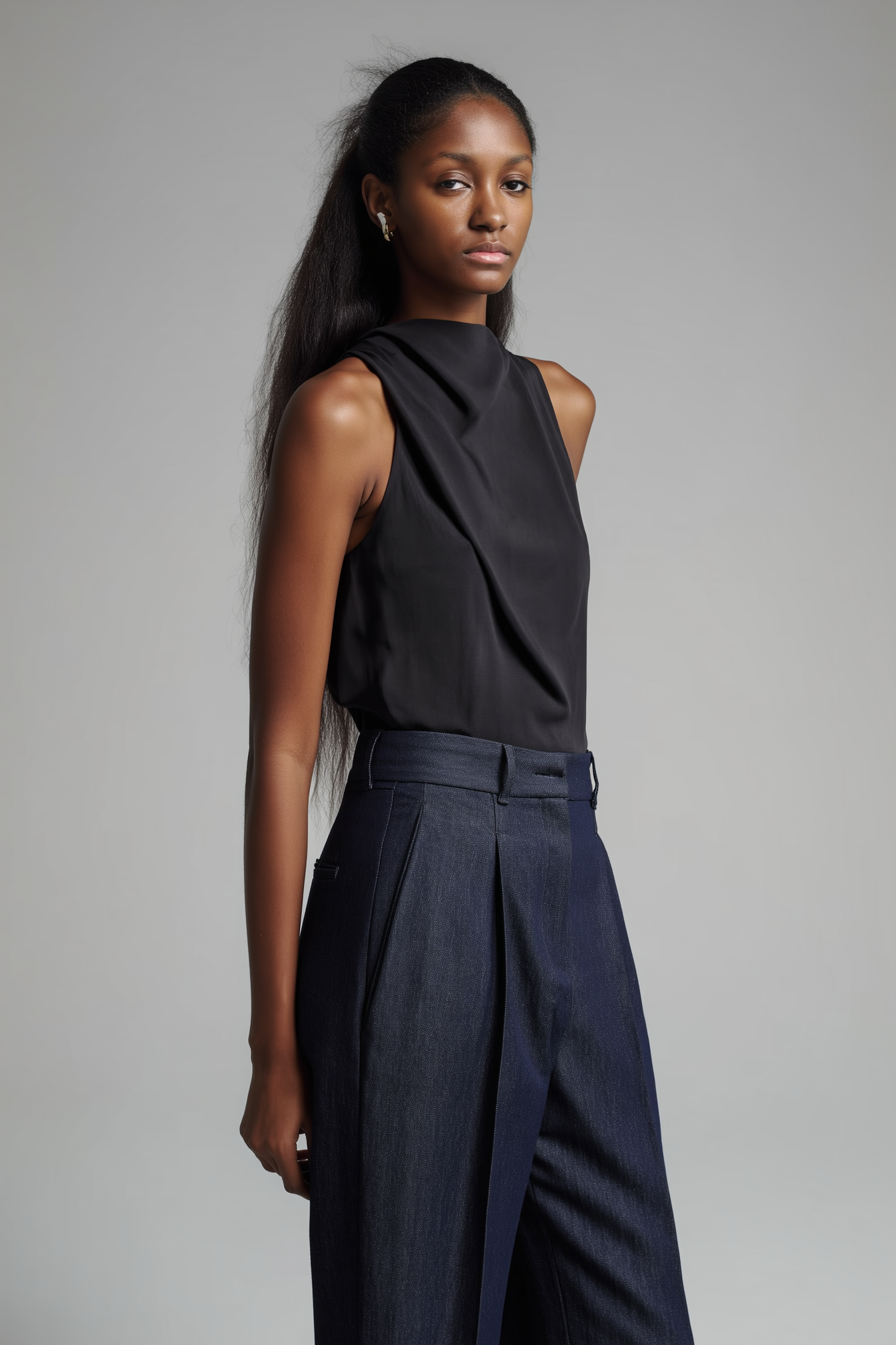 A young woman with long dark hair wearing a sleeveless black satin top and high-waisted dark blue trousers standing against a plain gray background.