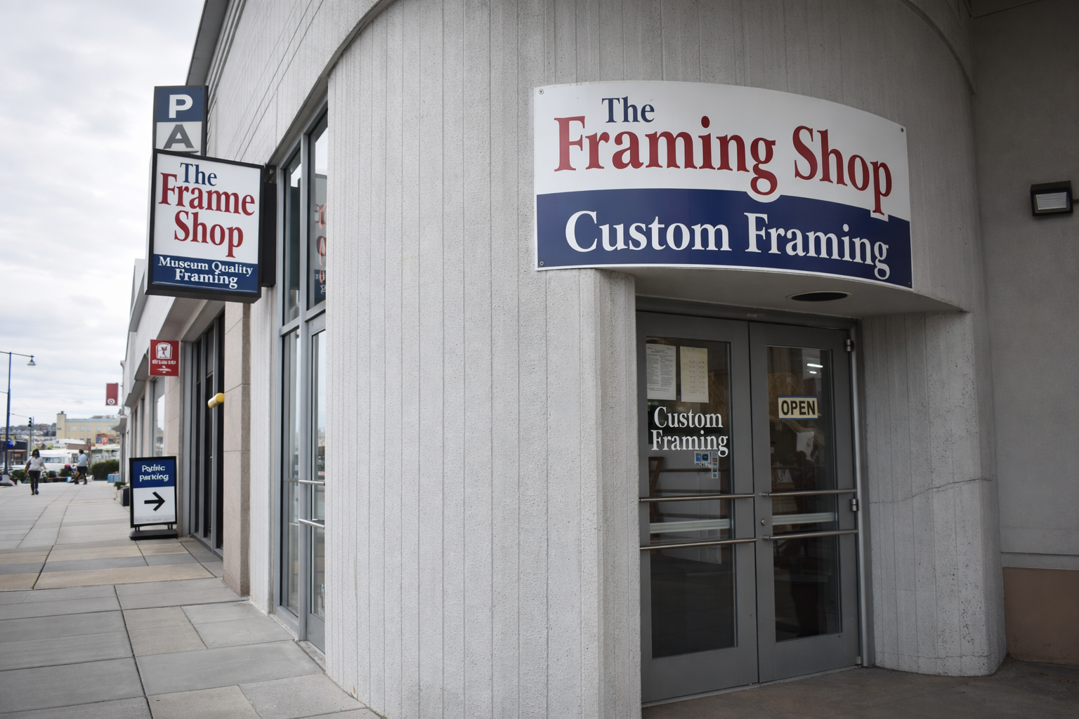 Exterior of a store called The Framing Shop with signs indicating custom framing, with additional signage on the sidewalk and nearby shops.