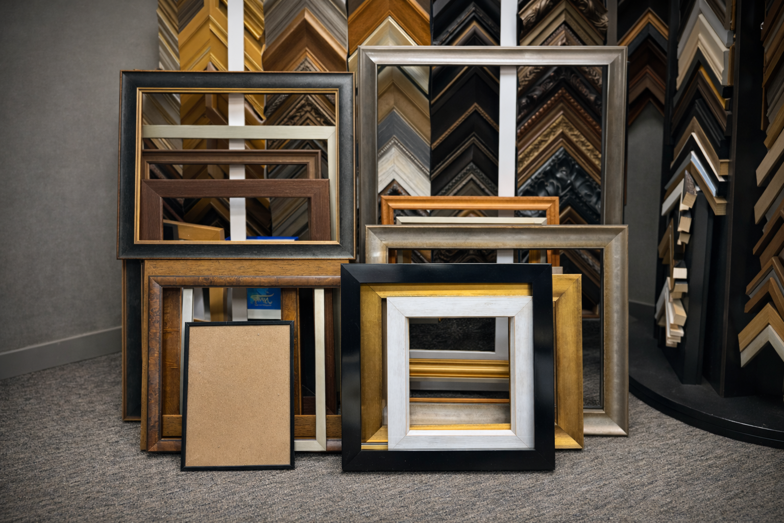 Display of various empty picture and photo frames in different sizes, colors, and designs, arranged on a carpeted floor against a backdrop of frame moldings and trim samples.