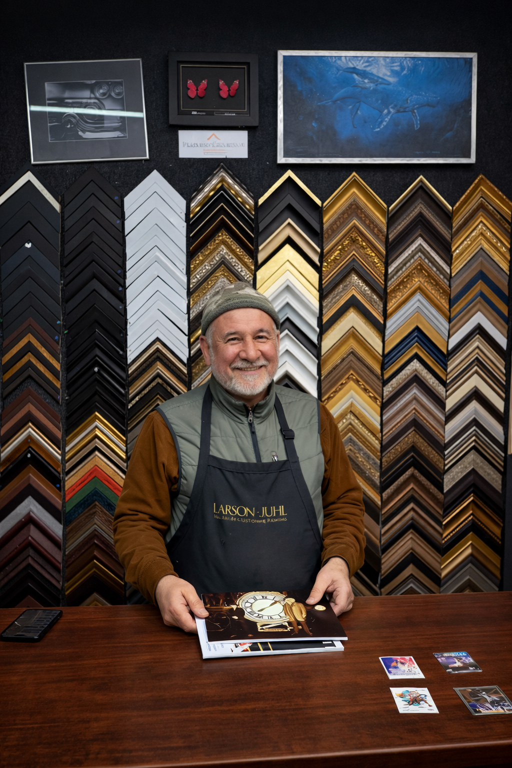 A smiling man with a beard, wearing a gray beanie. He is standing behind a wooden table with some artwork and cards for his framing project. Behind him is a wall displaying many sample frames and artwork.