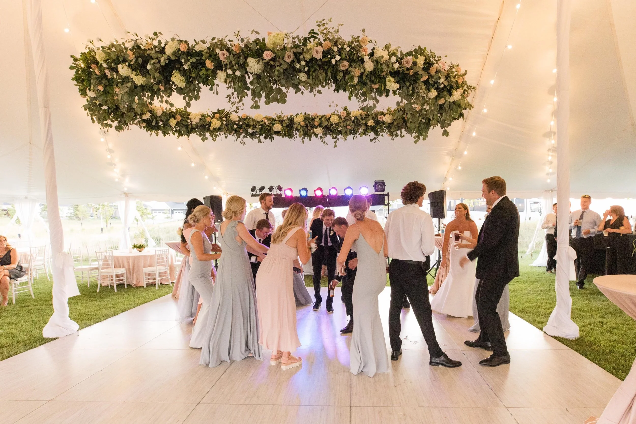 People dancing at a wedding reception inside a large decorated venue with floral ceiling arrangement and string lights.