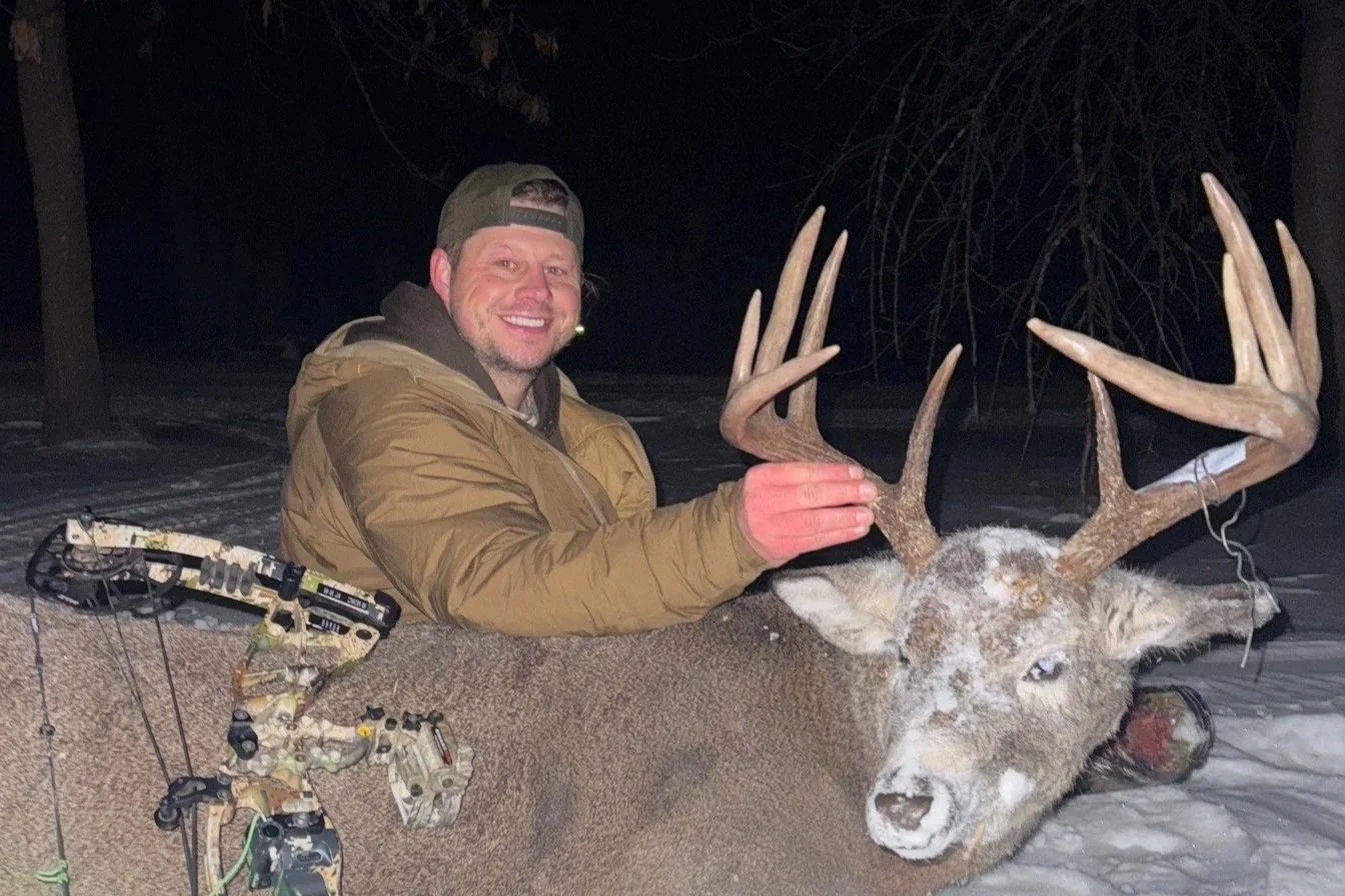 A man in a tan jacket and baseball cap smiling and holding the antlers of a large snow-covered deer in a snowy forest at night.