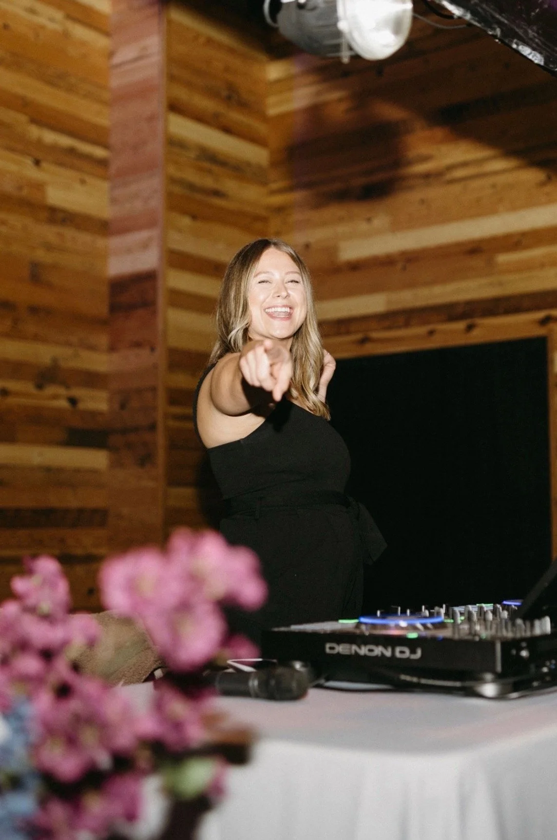 A young woman with long blonde hair wearing a black sleeveless top, smiling and pointing towards the camera, standing in front of a DJ mixer in a wooden-paneled room.