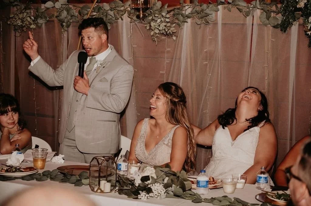A man in a light gray suit giving a speech at a wedding reception, with two women laughing and smiling as they sit at a decorated table. There are drinks, greenery, and decorations on the table and background.