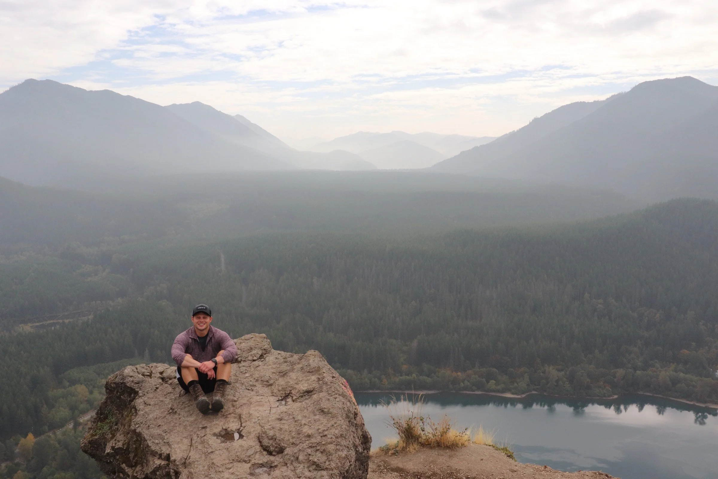 A young man with a cap and a gray jacket sitting on a large rock atop a mountain overlooking a forest and a lake with distant mountains in the background on a cloudy day.