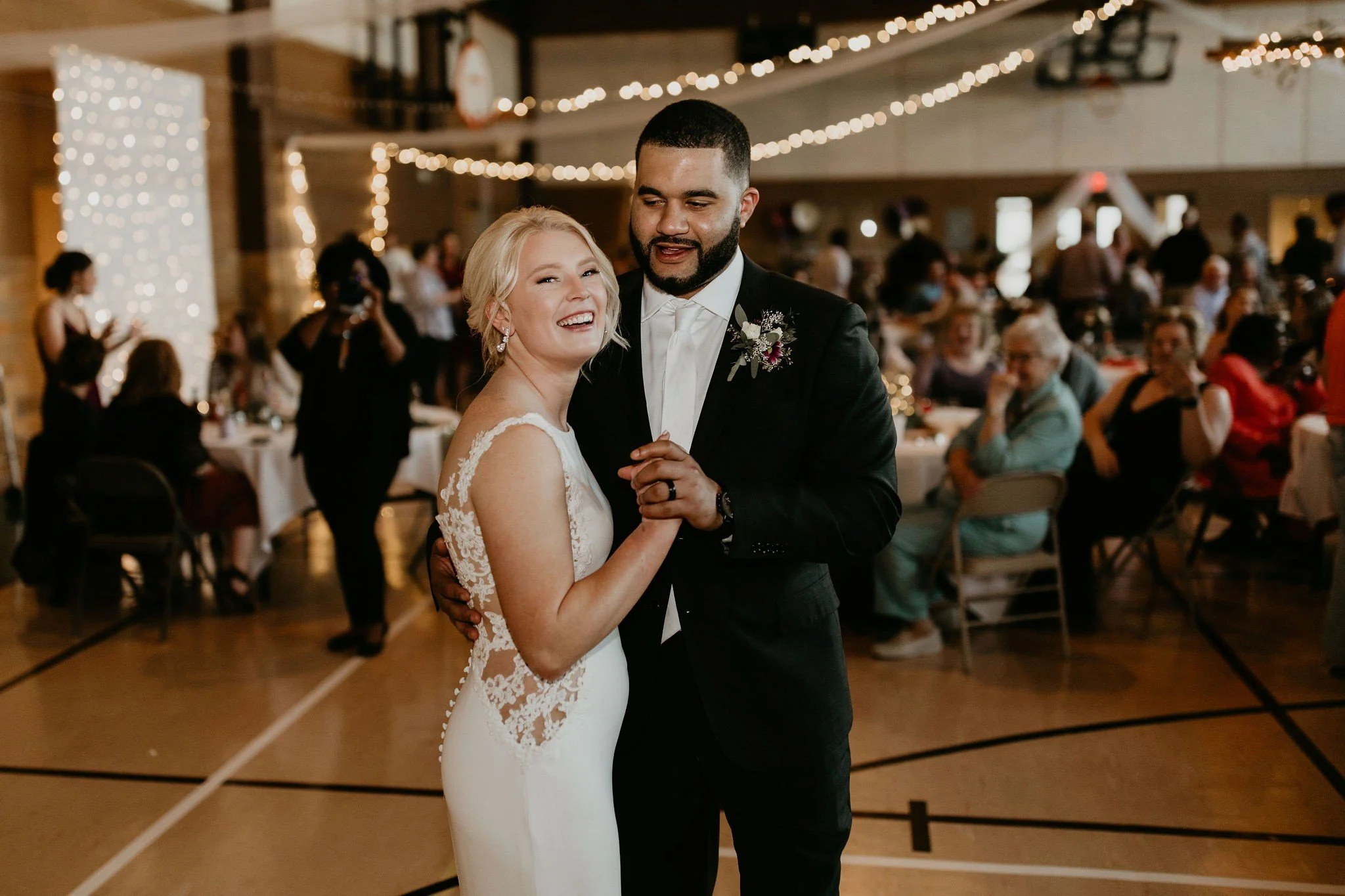 A bride and groom dance together at a wedding reception inside a decorated hall, with guests seated at tables around them.