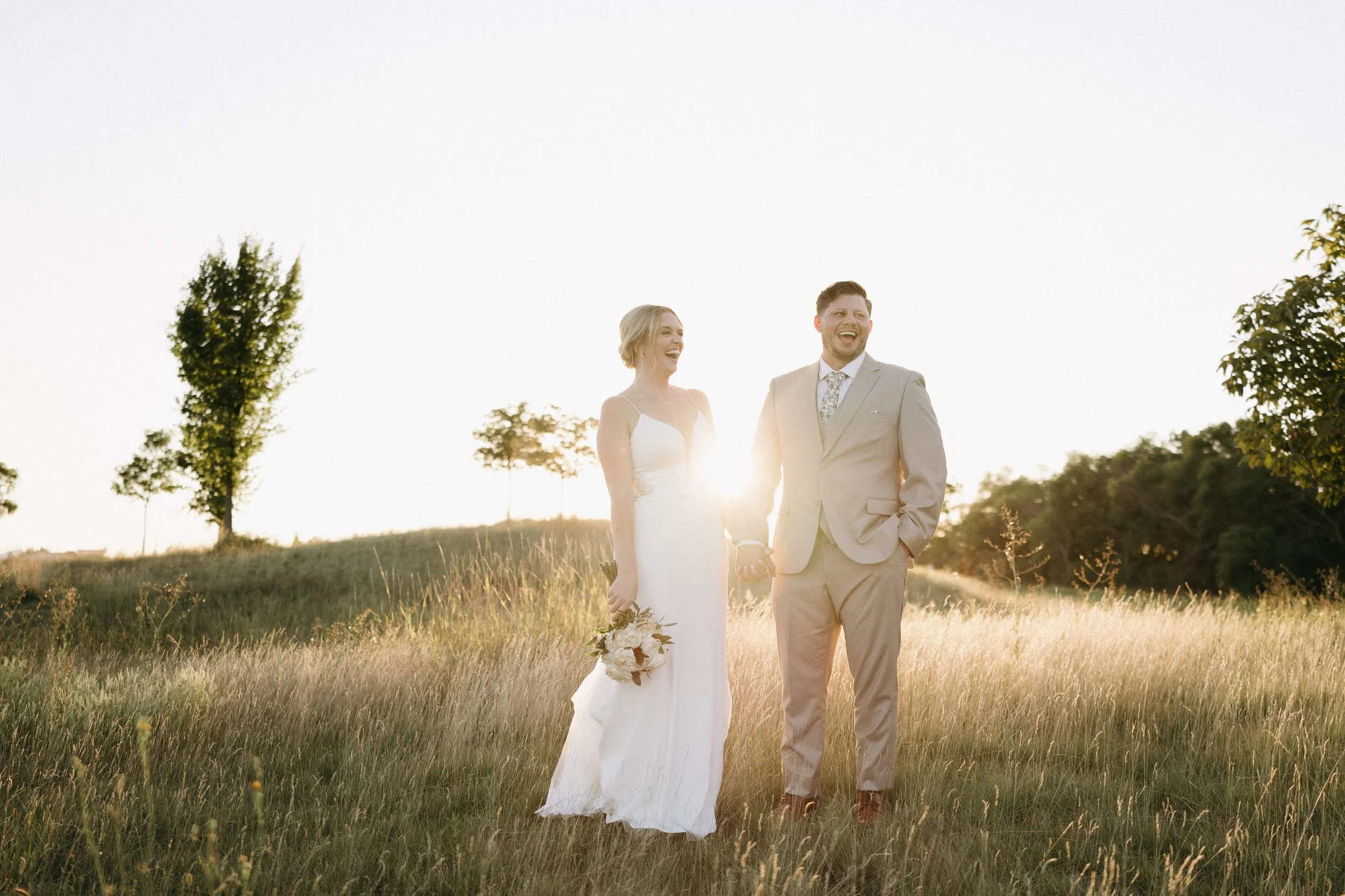 A couple dressed in wedding attire holding hands and smiling in a field at sunset, with trees in the background.