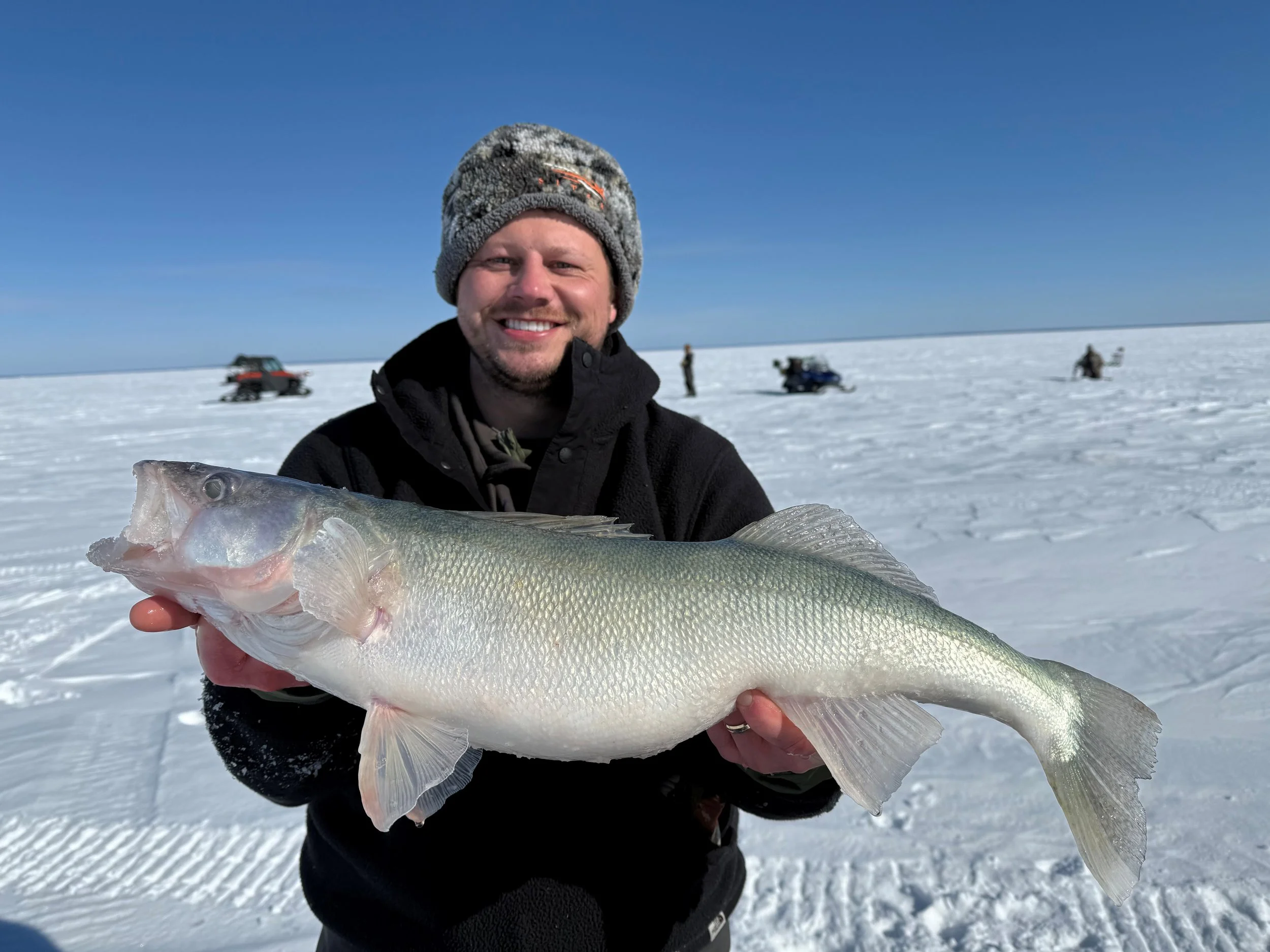 A man smiling and holding a large fish on a snowy landscape with several snowmobiles and people in the background under a clear blue sky.