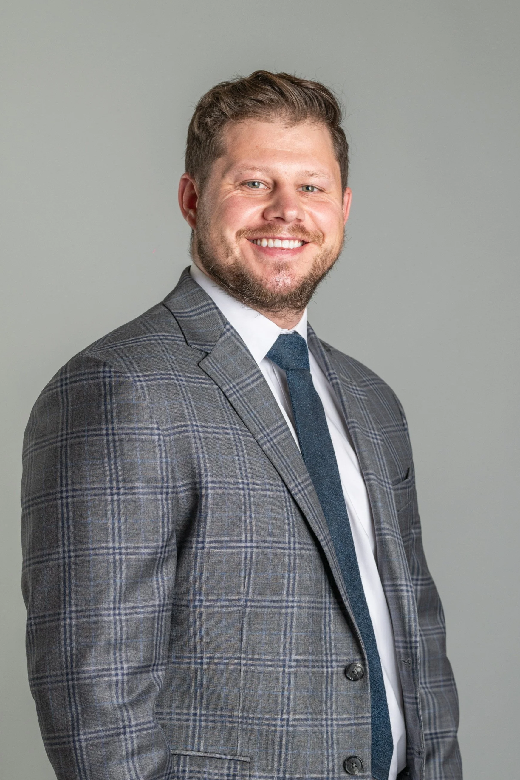 A smiling man with short hair, beard, and mustache, wearing a gray plaid suit, white shirt, and dark blue tie, standing against a plain gray background.