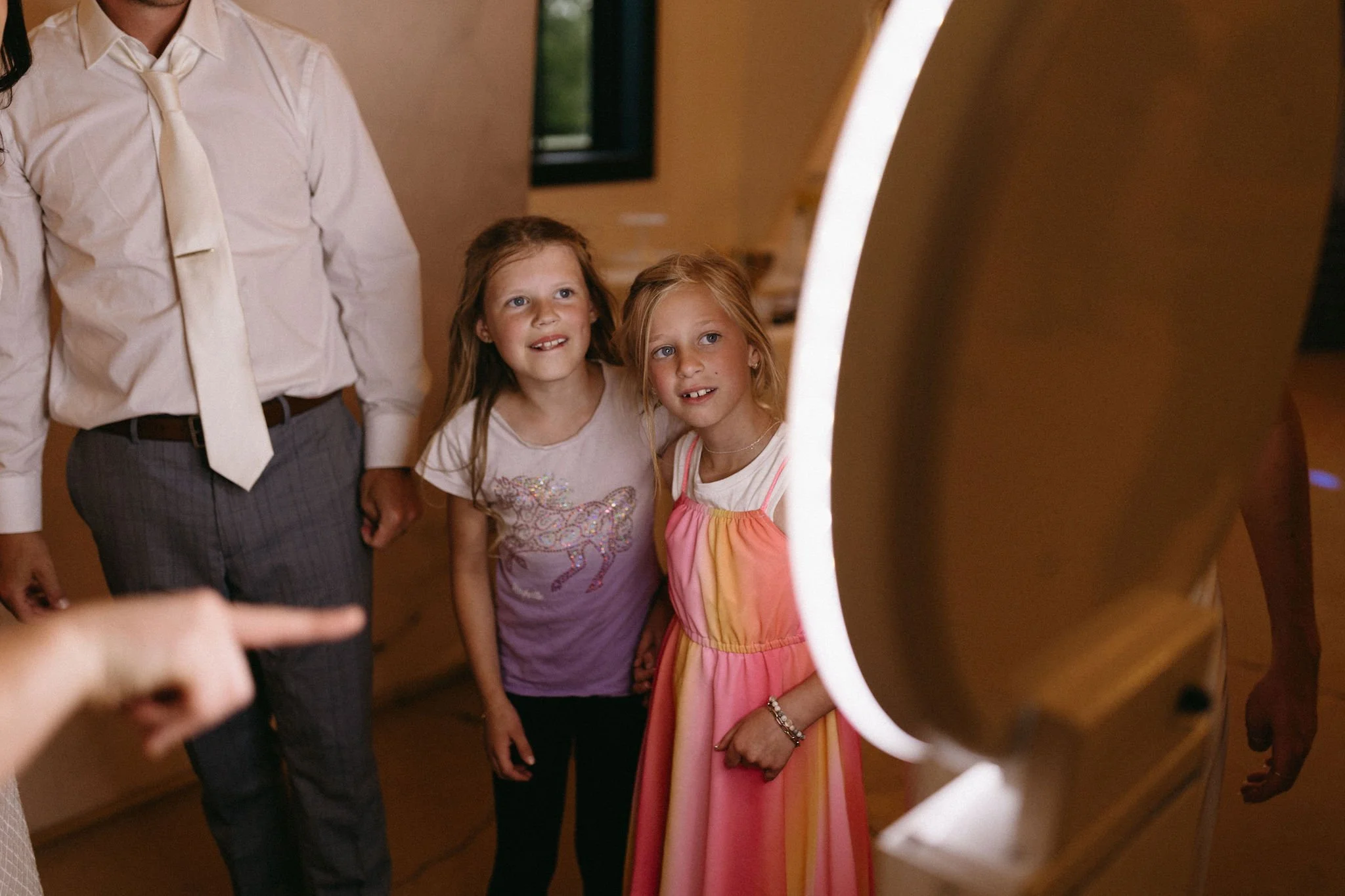 Two young girls admiring an object or display inside a circular frame, with adults nearby in a warmly lit indoor setting.