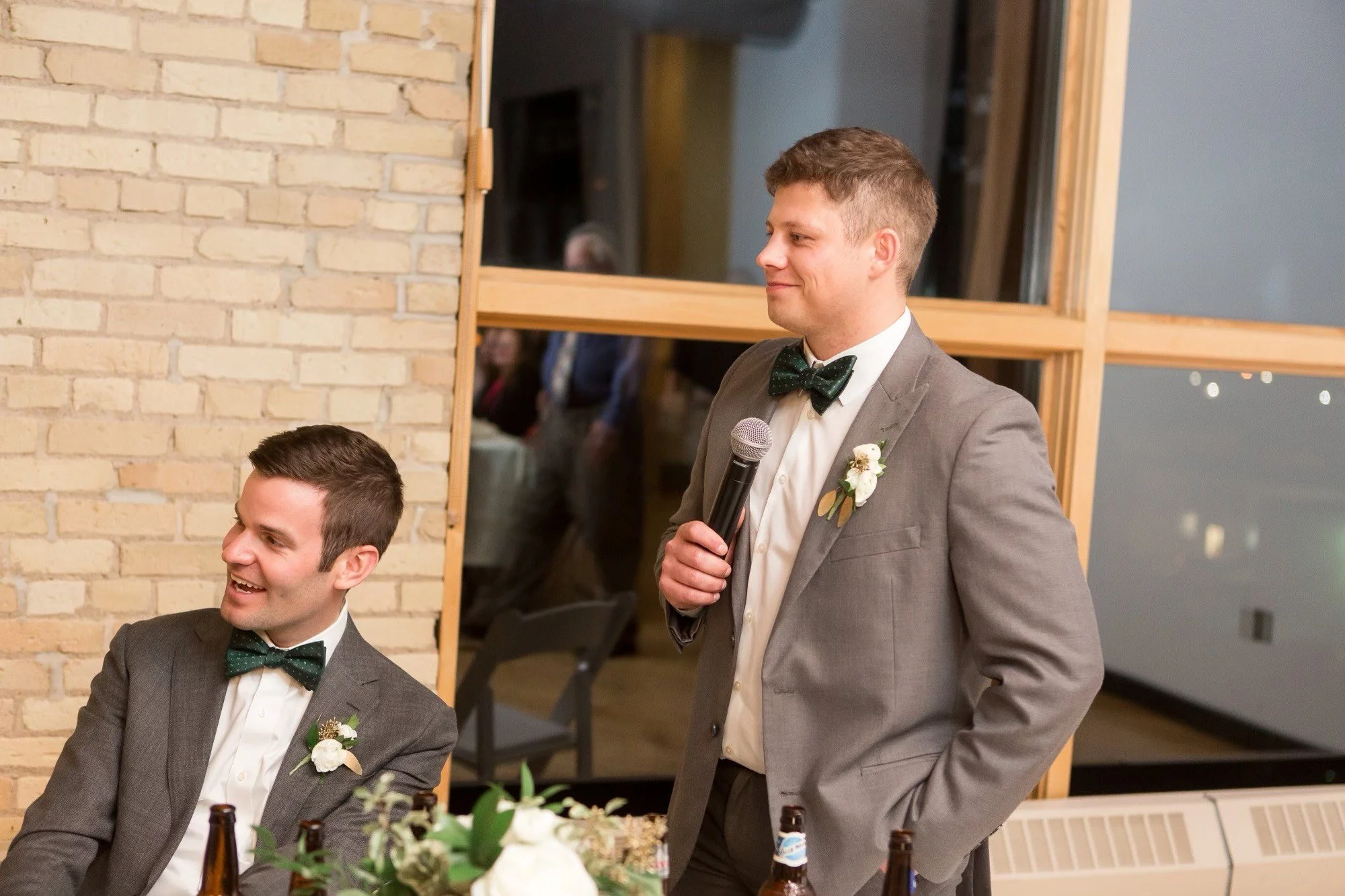 A man in a gray suit with a bow tie and boutonniere is standing and speaking into a microphone at a social event. Two men in suits with bow ties are seated at a table in front of him, smiling. The background shows a large window and a brick wall.