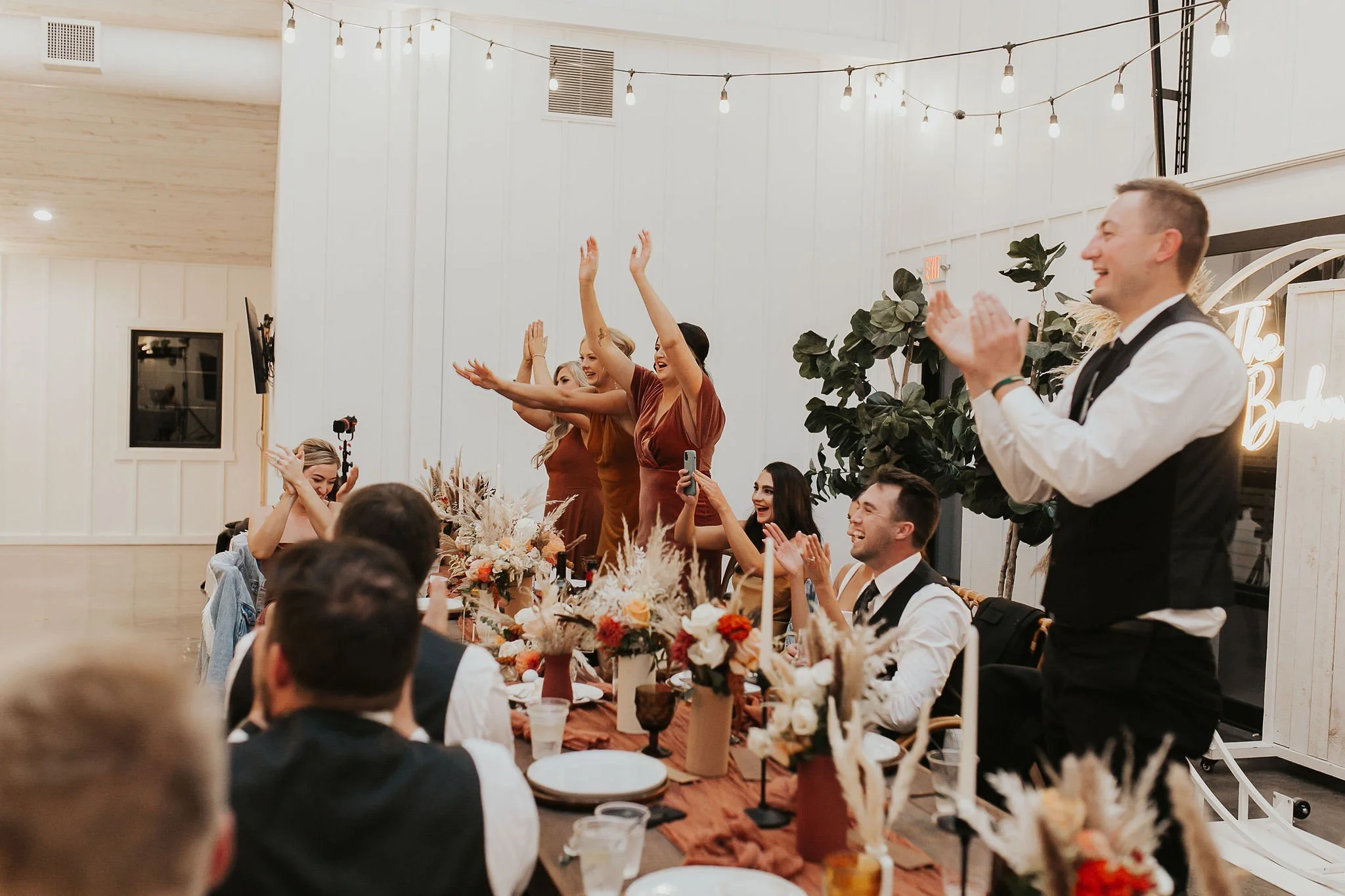 A group of people celebrating at a wedding reception, with women standing and raising their hands in joy, and men seated at a decorated table with floral centerpieces. The setting is indoors with white walls and string lights overhead.