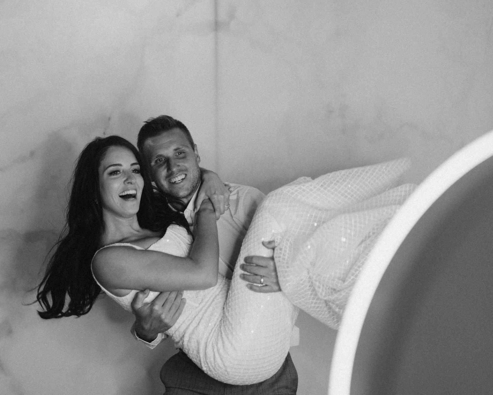 A couple on a bed, with the man lying down and the woman sitting on his lap, both smiling and looking at the camera. The bed has a patterned blanket, and the background is plain.