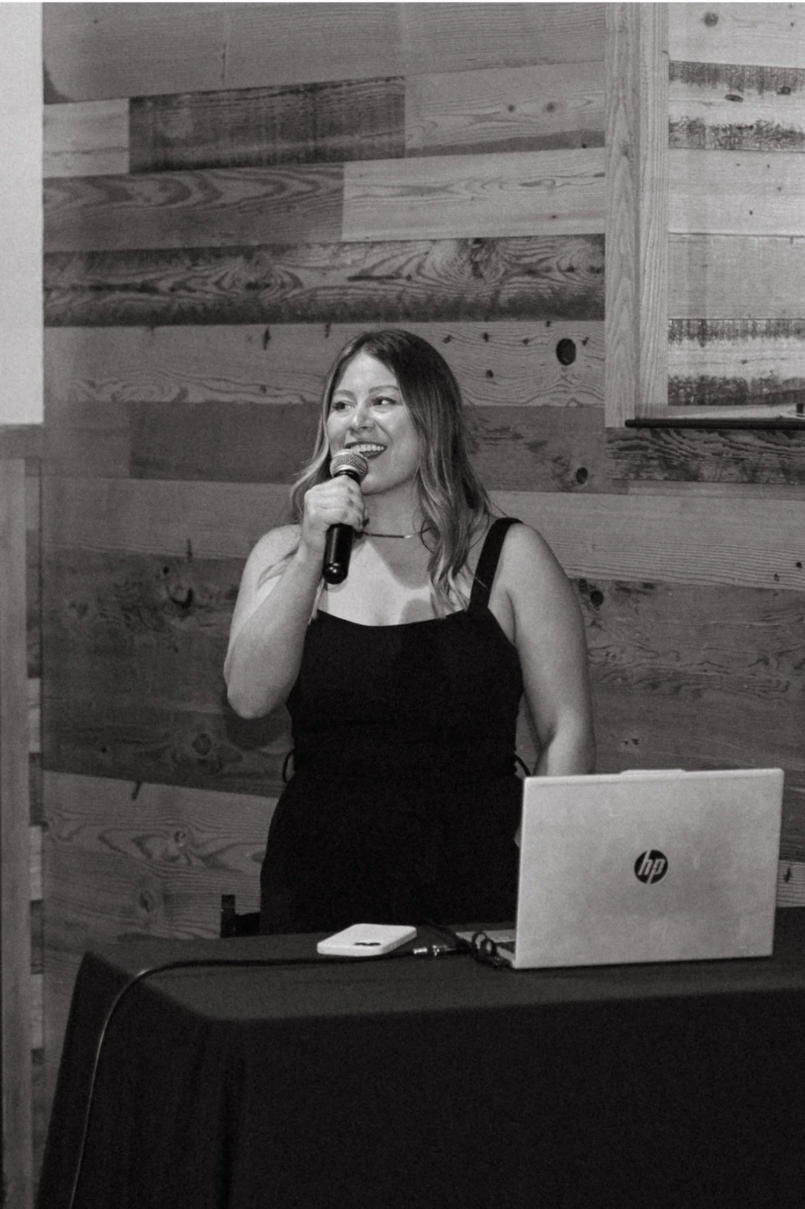 Black and white photo of a woman holding a microphone, standing in front of a wooden wall, with an HP laptop and a smartphone on a table in front of her.