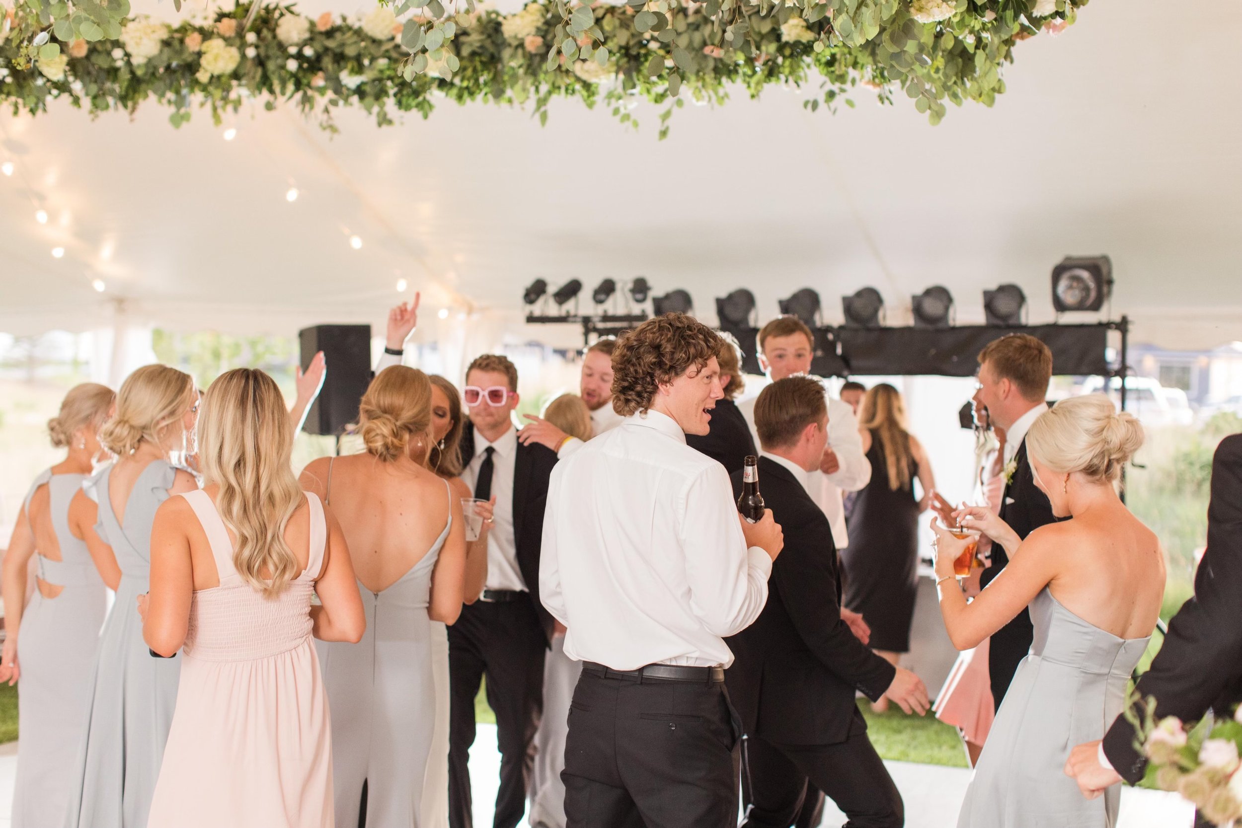 People dancing and socializing at a wedding reception under a decorated tent with floral arrangements and string lights.