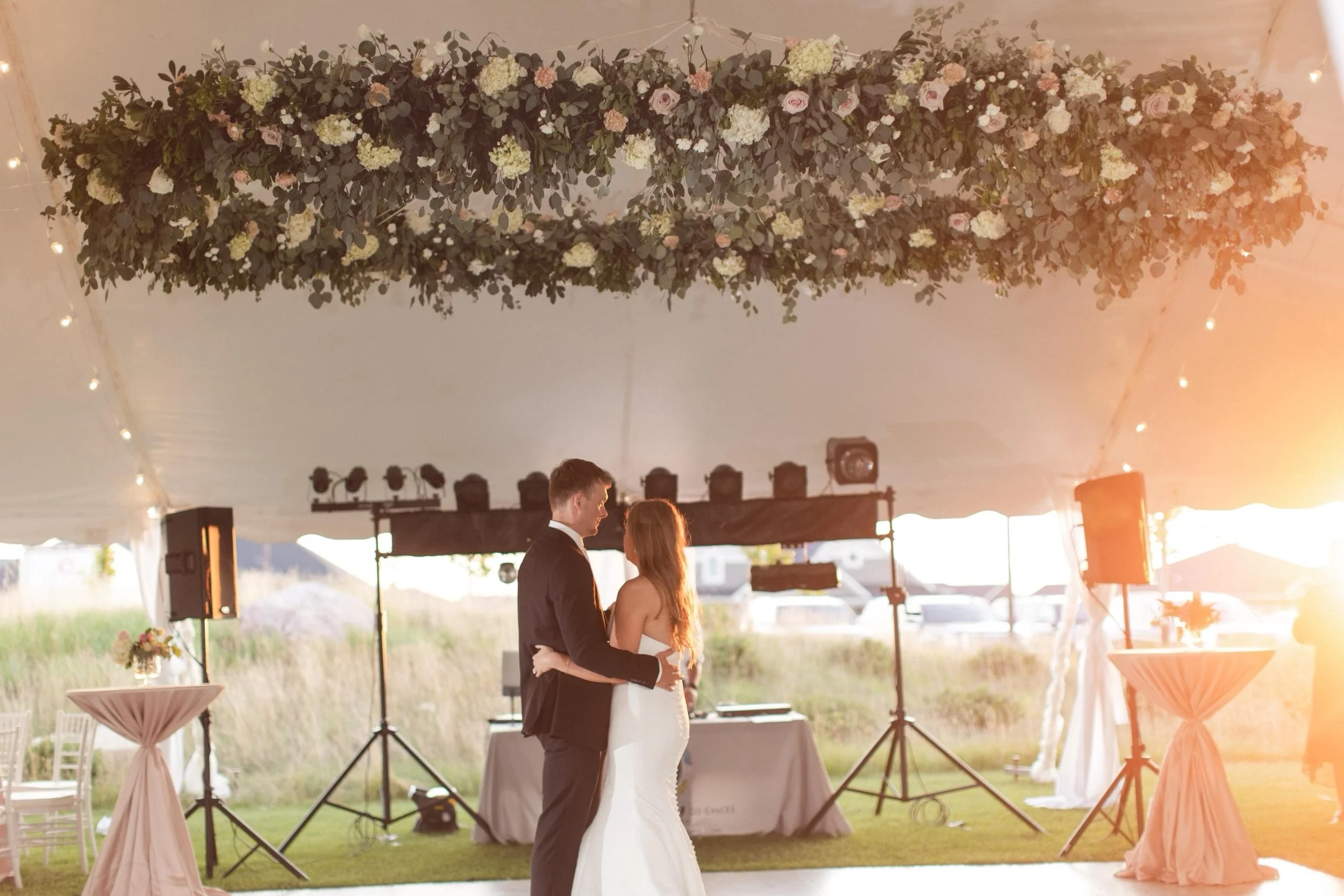 A bride and groom sharing a kiss during their wedding reception under a decorated tent with floral arrangements hanging from the ceiling.