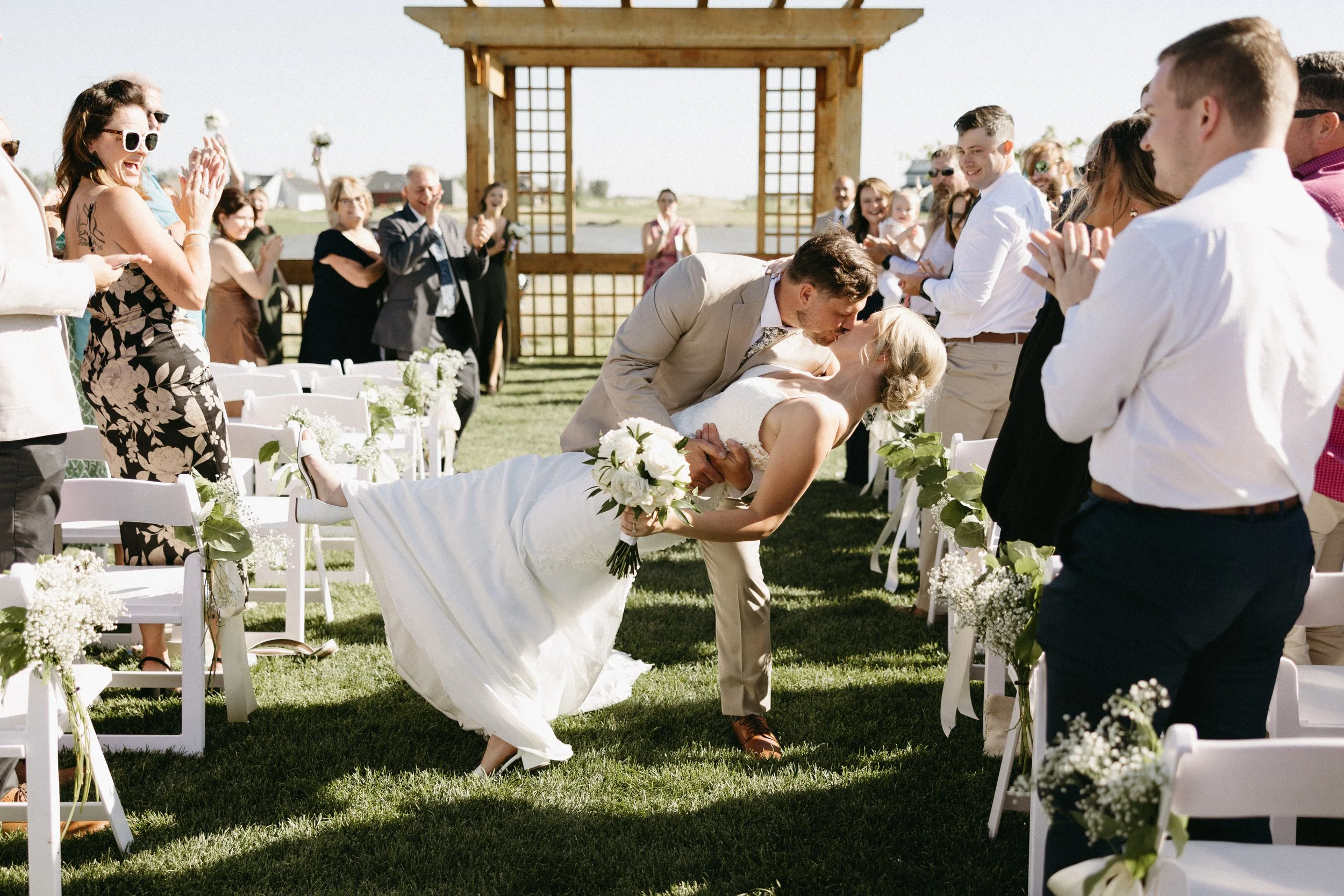 A bride and groom kissing during their wedding ceremony outdoors, with guests clapping and smiling around them, on a sunny day with a wooden arch and green grass.