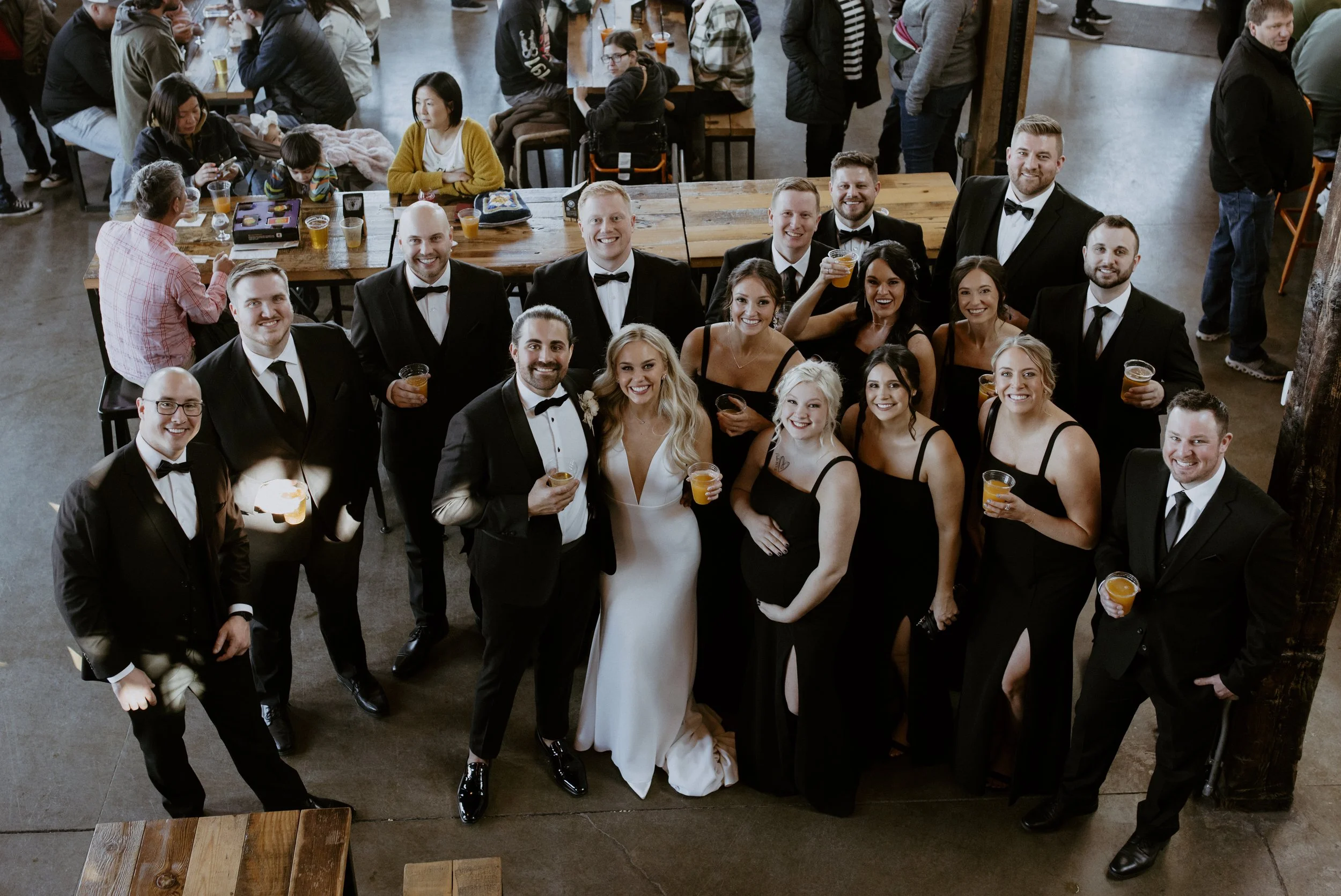Group of people dressed in formal attire, including tuxedos and wedding dresses, celebrating at a wedding reception, with some holding drinks.