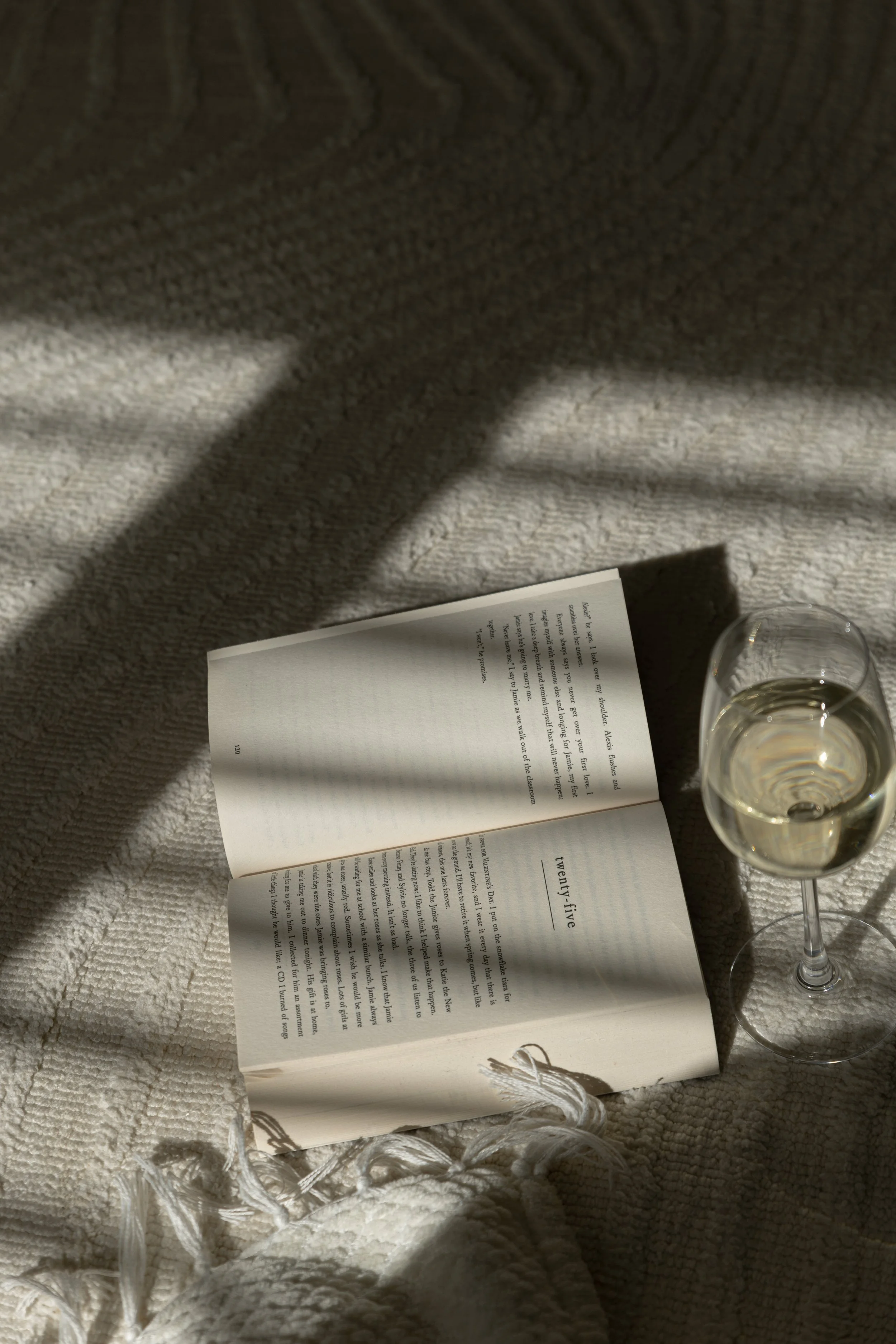 An open book with text and the number twenty-five on one page, placed on a textured, light-colored blanket. Next to the book is a glass of white wine. Sunlight creates shadows on the scene.