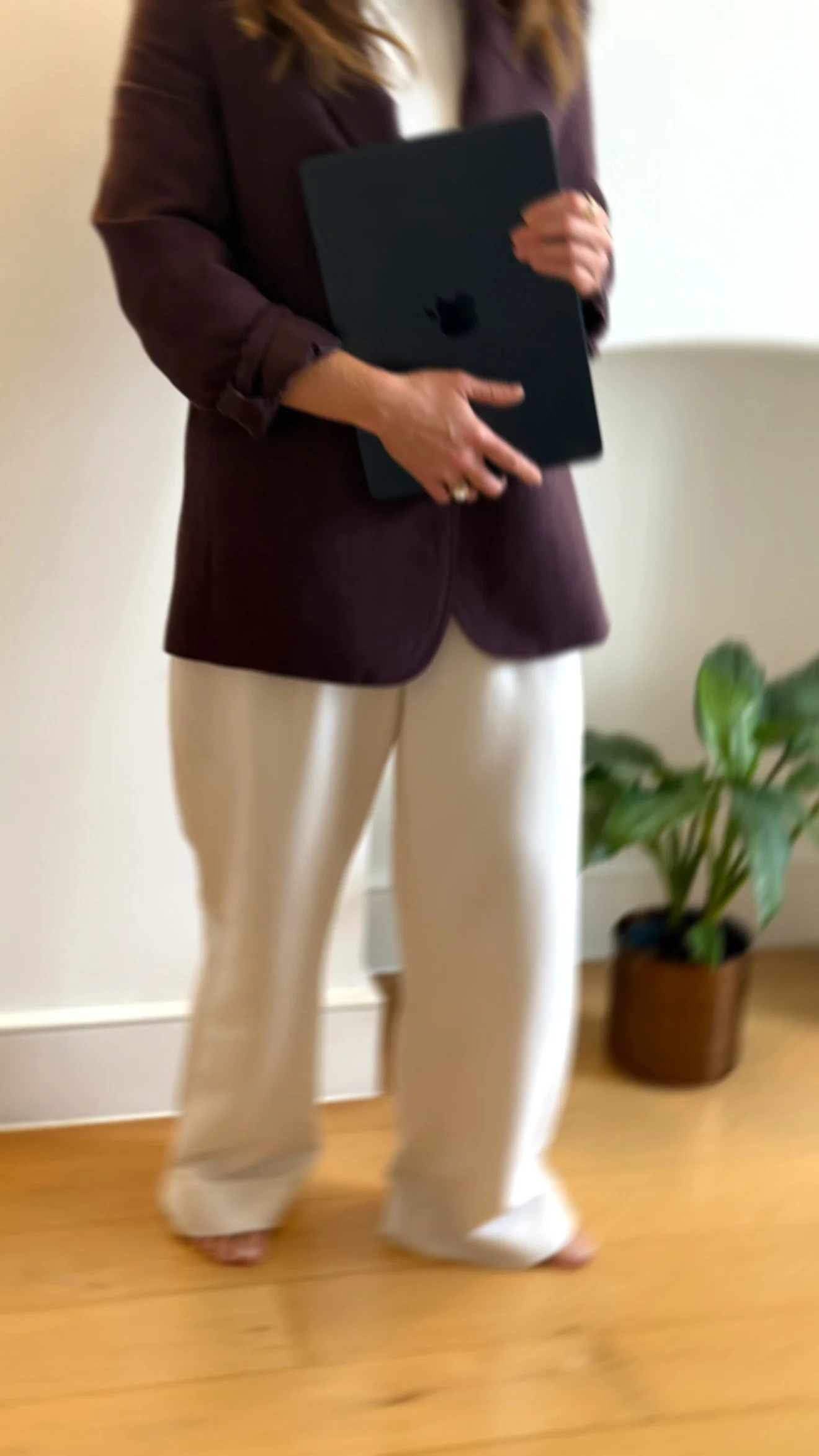 A woman dressed in a brown blazer and cream-colored pants holding a black laptop standing indoors near a potted plant.