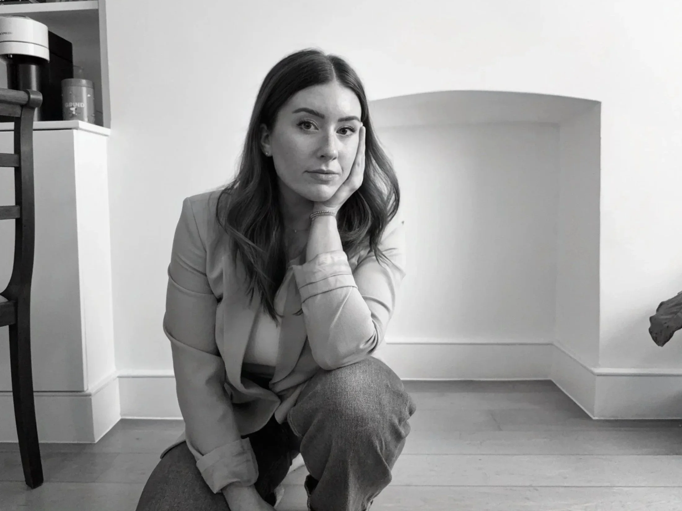 A black-and-white photo of a woman with wavy hair, squatting on the floor with her hand resting on her cheek, looking directly at the camera in a room with minimal decor.