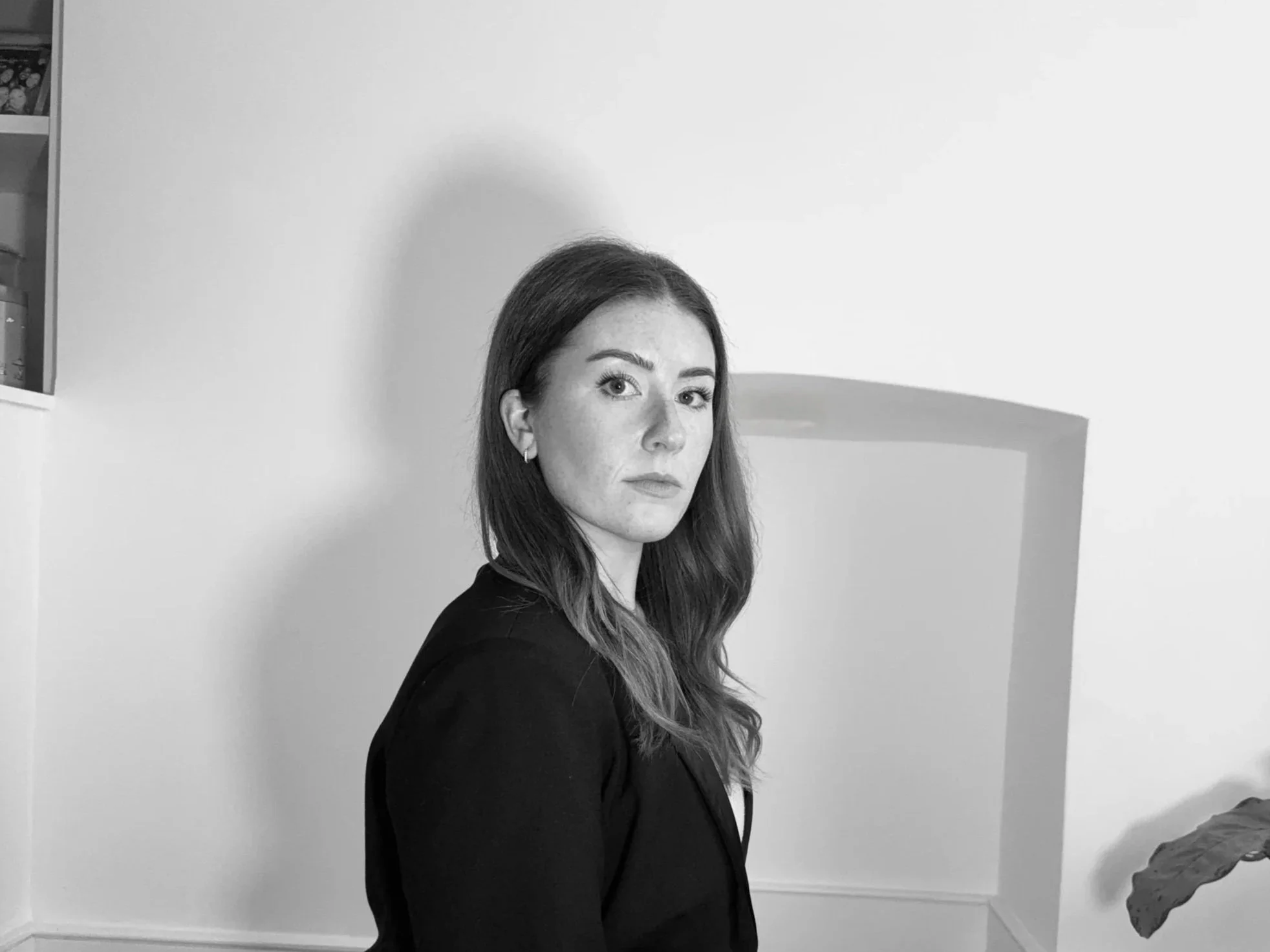 Black and white photo of a woman with long hair wearing a dark blazer, standing in front of a plain wall.