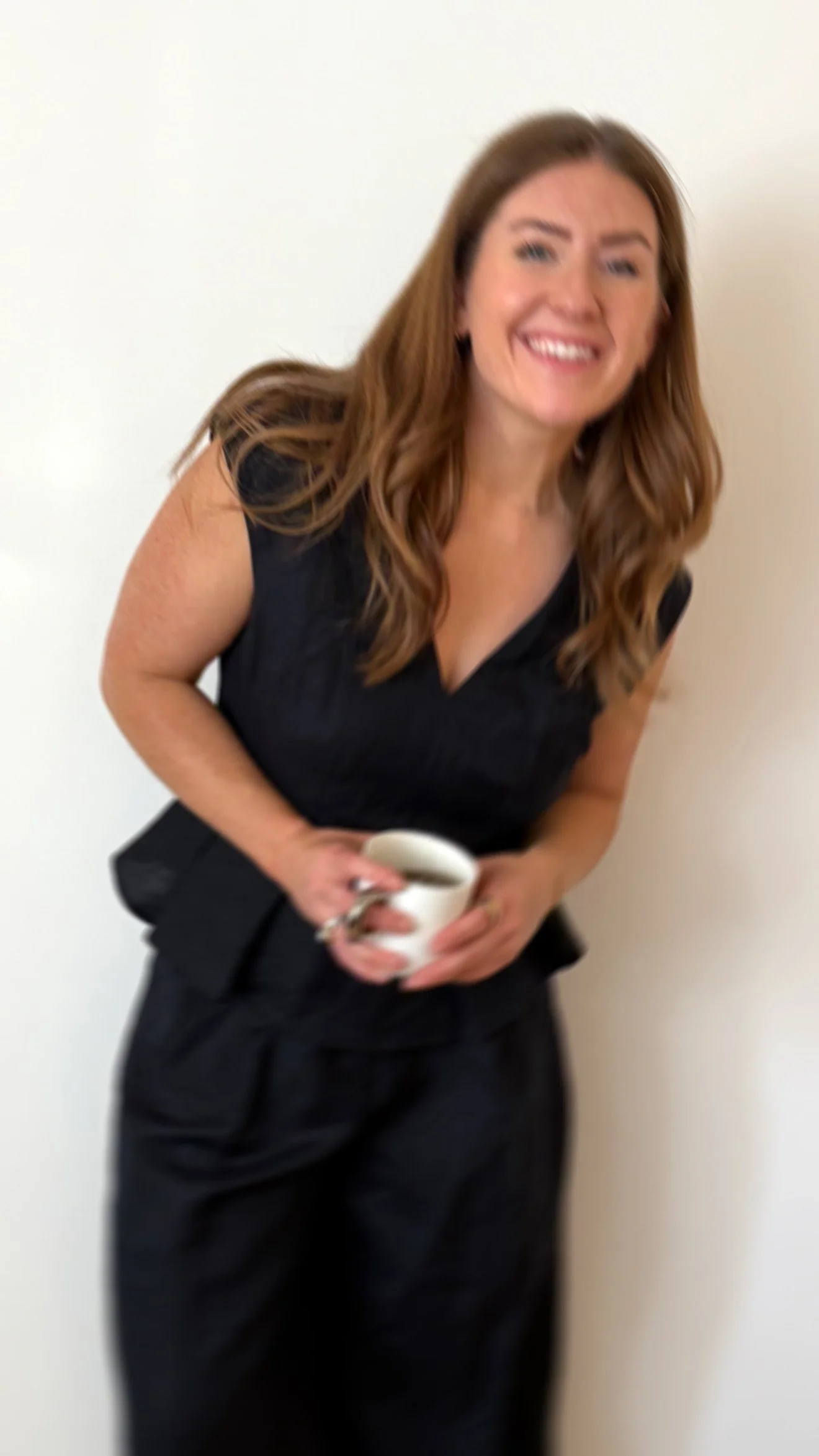 A smiling woman with long, wavy brown hair wearing a black dress holding a white coffee mug.